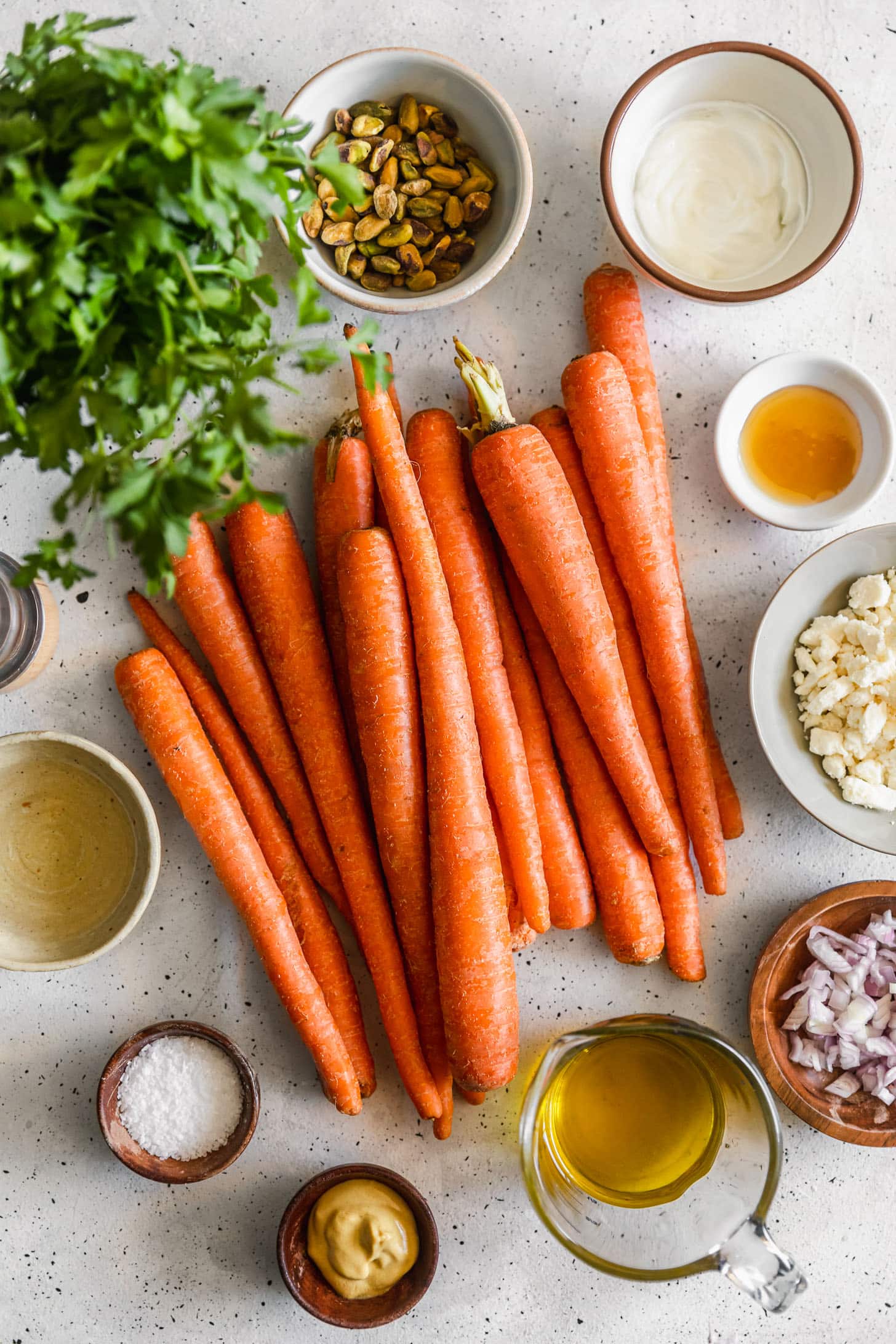 Carrots, a white bowl of feta, a white bowl of pistachios, a wood bowl of shallots, parsley, and white bowls of oil, honey, vinegar, and mustard on a white counter.