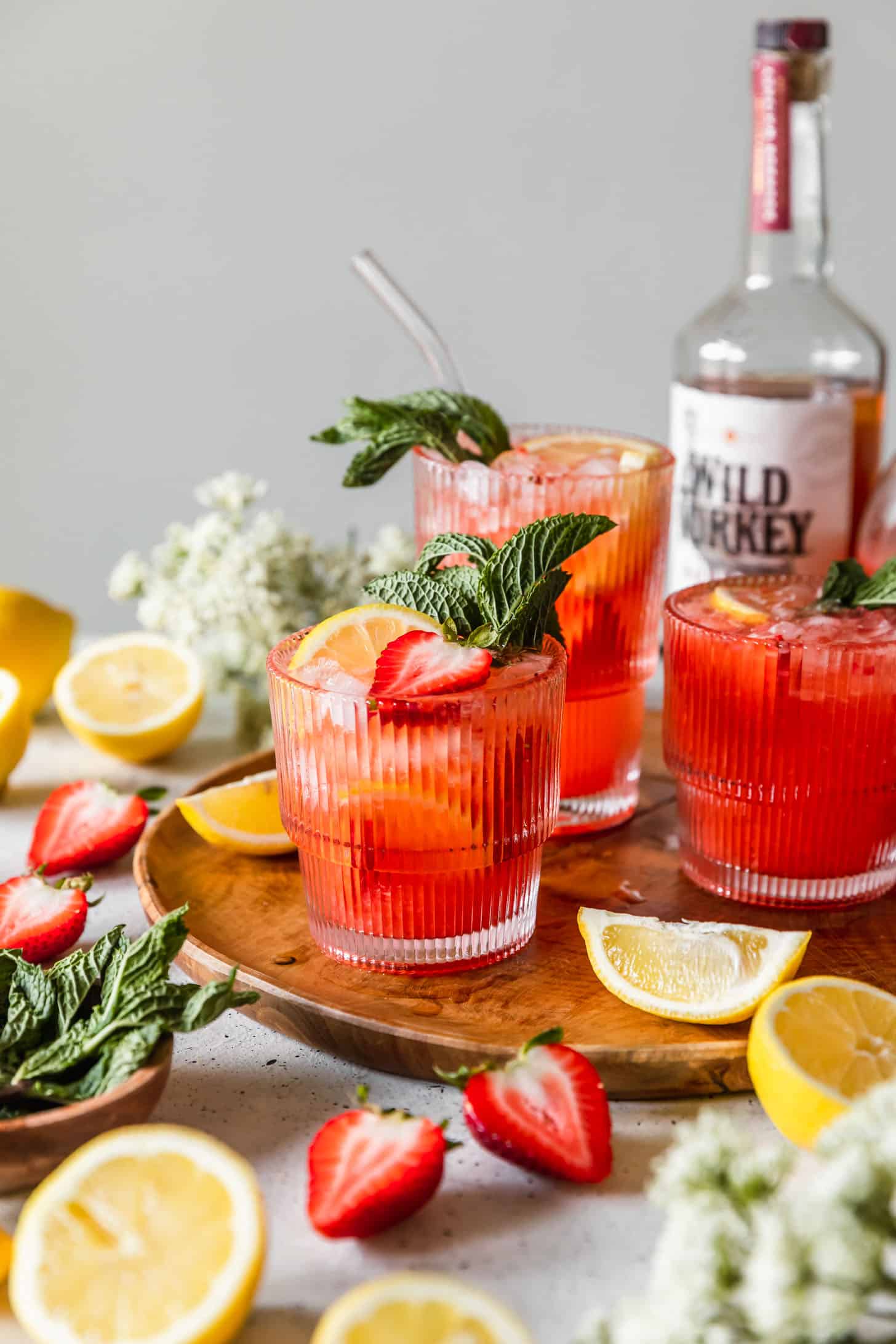 Three glasses of strawberry lemonade cocktail next to lemons, strawberries, mint, white flowers, and a bottle of bourbon on a white background.