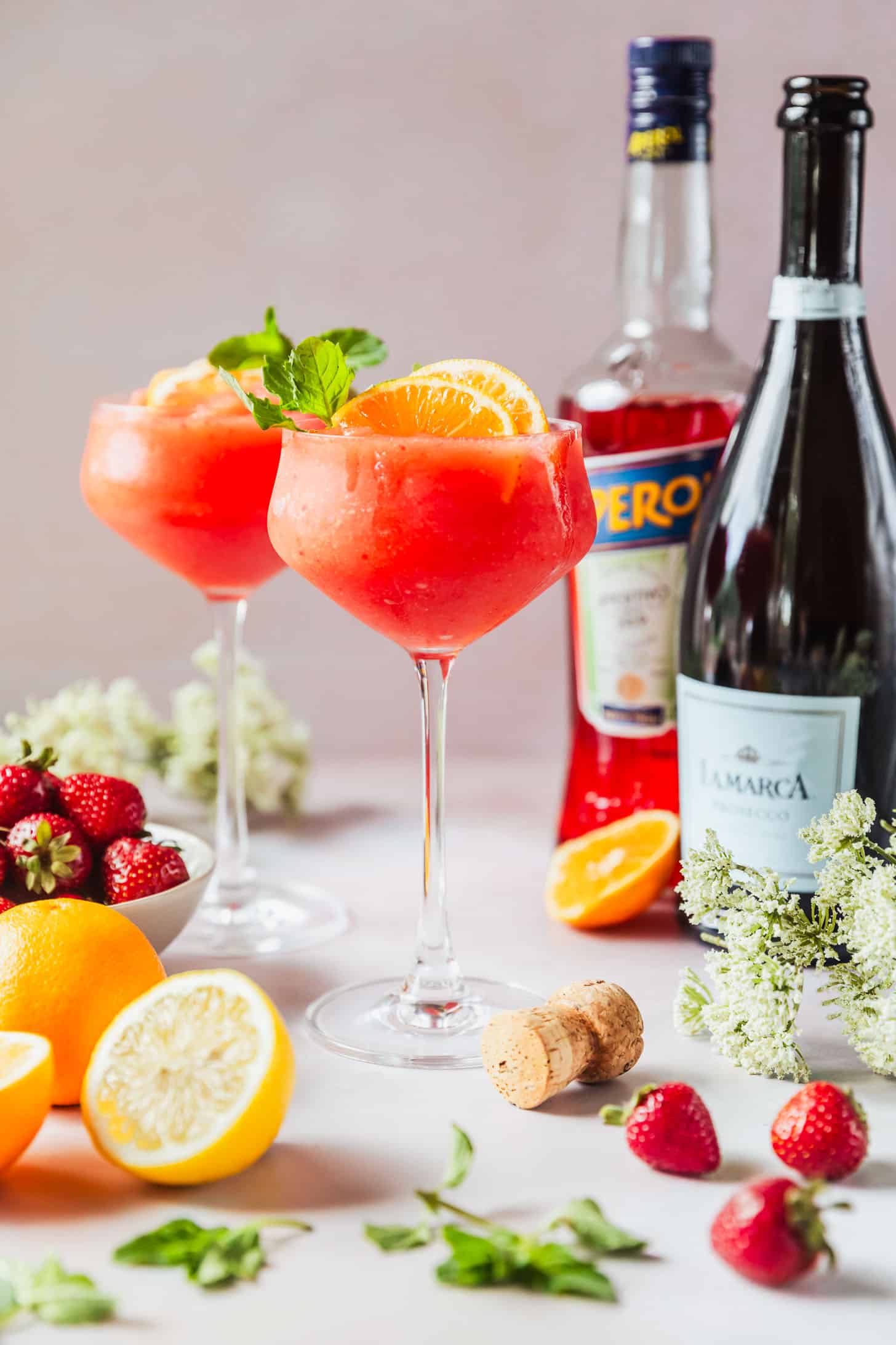 Two frozen Aperol spritz cocktails in coupe glasses on a pink background next to a white bowl of strawberries, lemons, oranges, white flowers, a bottle of Prosecco, and a bottle of Aperol.