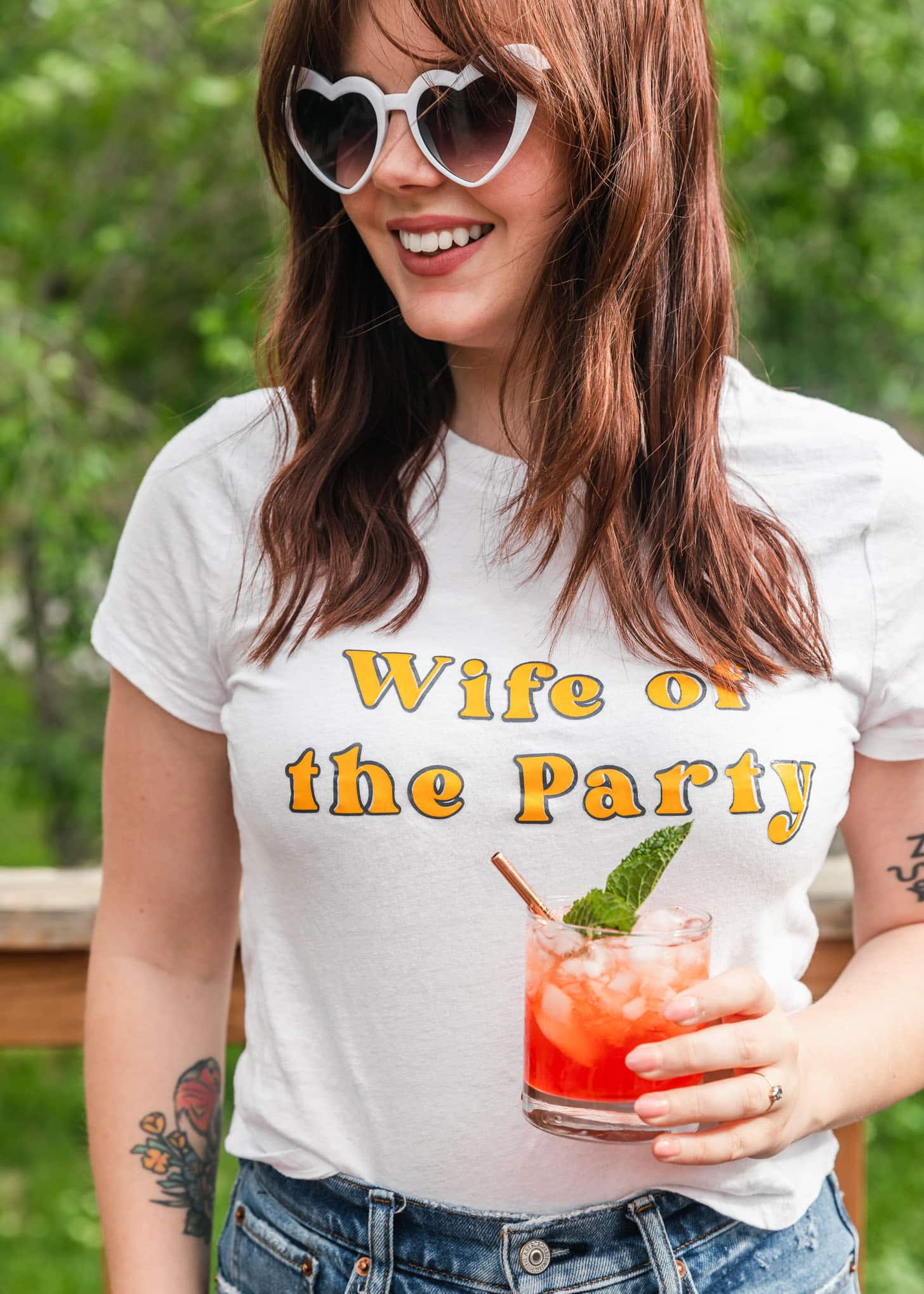 A woman with wavy dark red hair and bangs, white heart sunglasses, and a white "Wife of the Party" t-shirt holding a pink cocktail in front of trees.