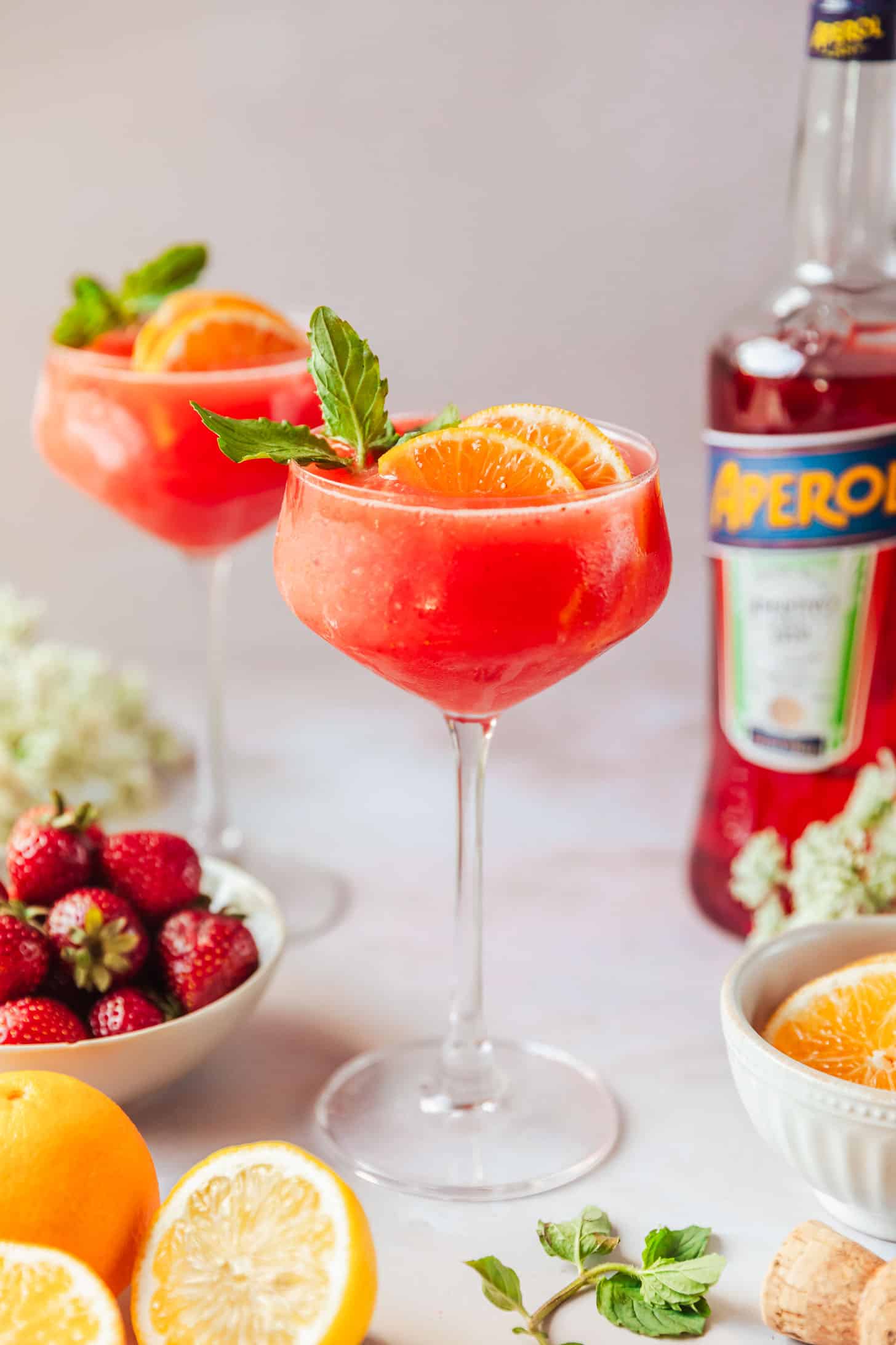 Two frozen Aperol spritz cocktails on a pink counter next to a white bowl of strawberries, a bottle of Aperol, white flowers, a white bowl of orange slices, and lemons.