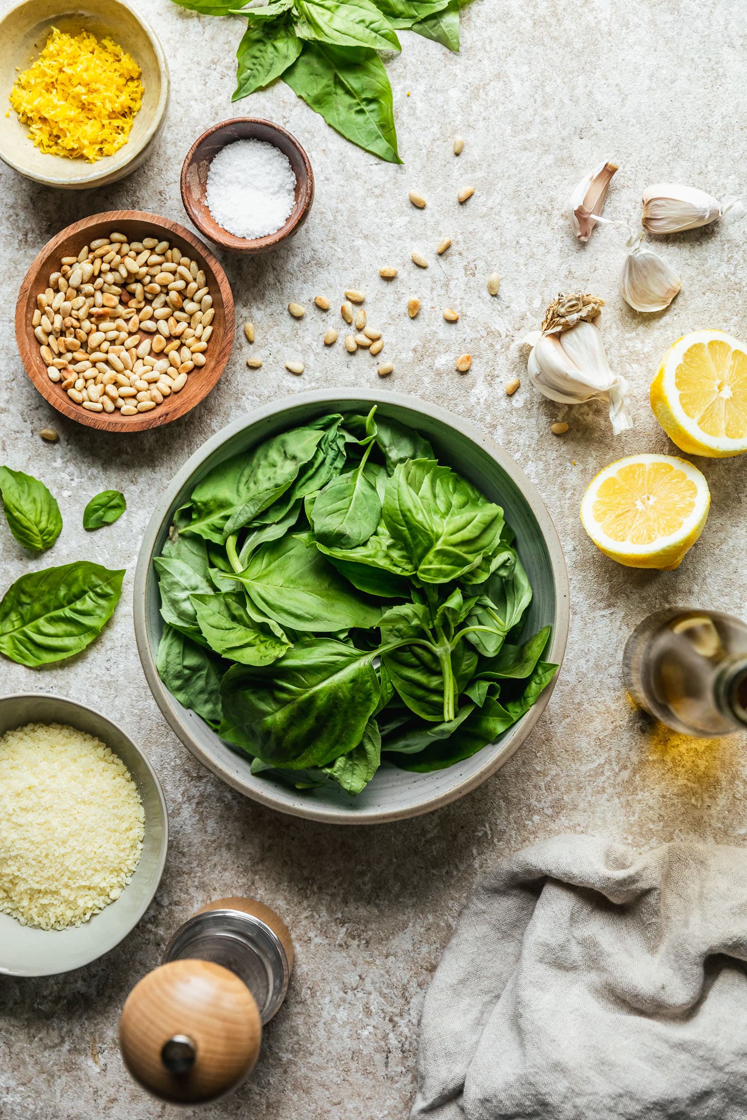 A grey bowl of basil on a beige counter next to halved lemons, a jar of olive oil, a beige linen, a grey bowl of Parmesan, a white bowl of lemon zest, and a wood bowl of pine nuts.