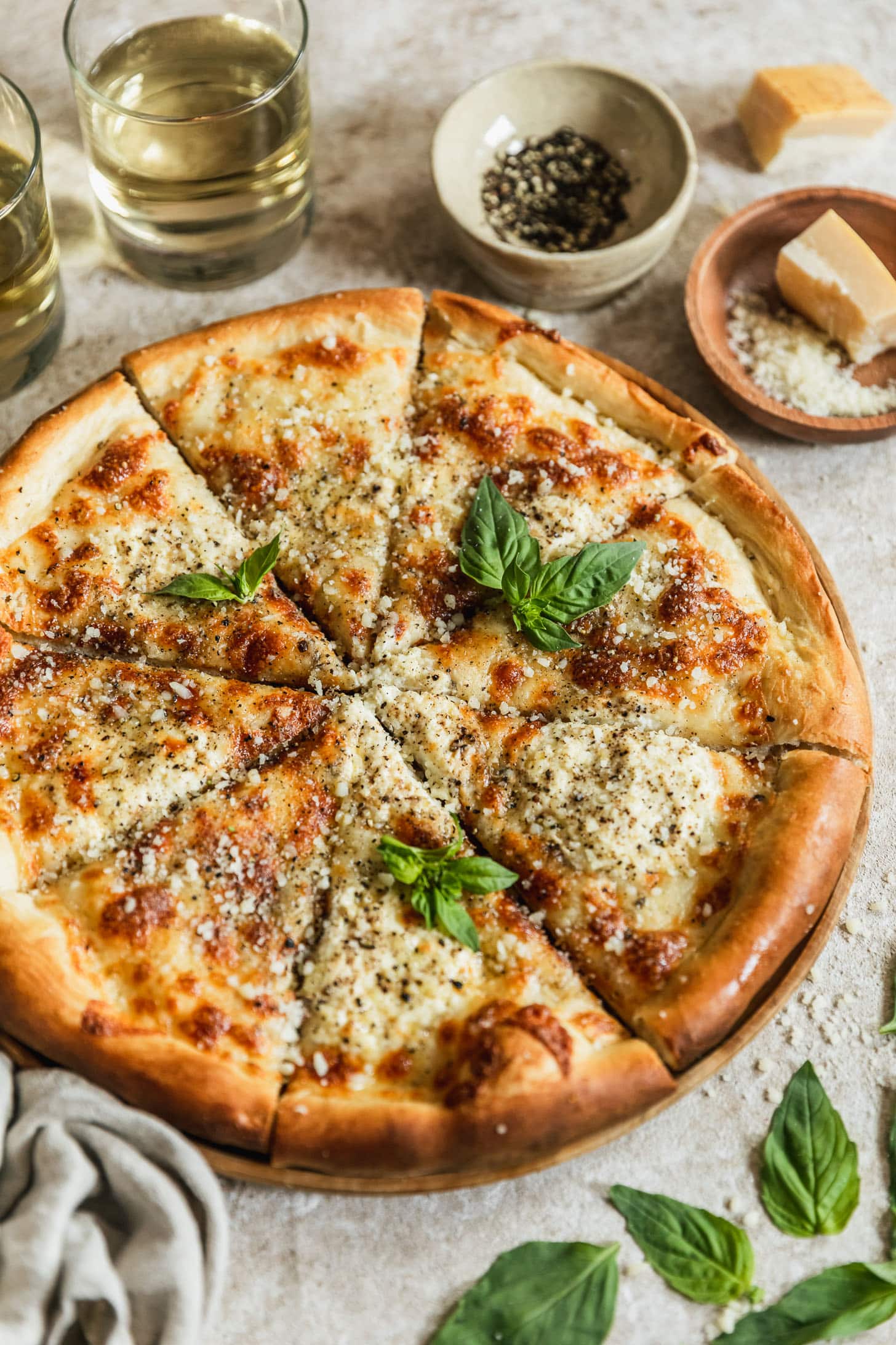 A wood board with a cacio e pepe pizza on a beige counter next to fresh basil, two glasses of white wine, a white bowl of pepper, and a wood bowl of Pecorino Romano.