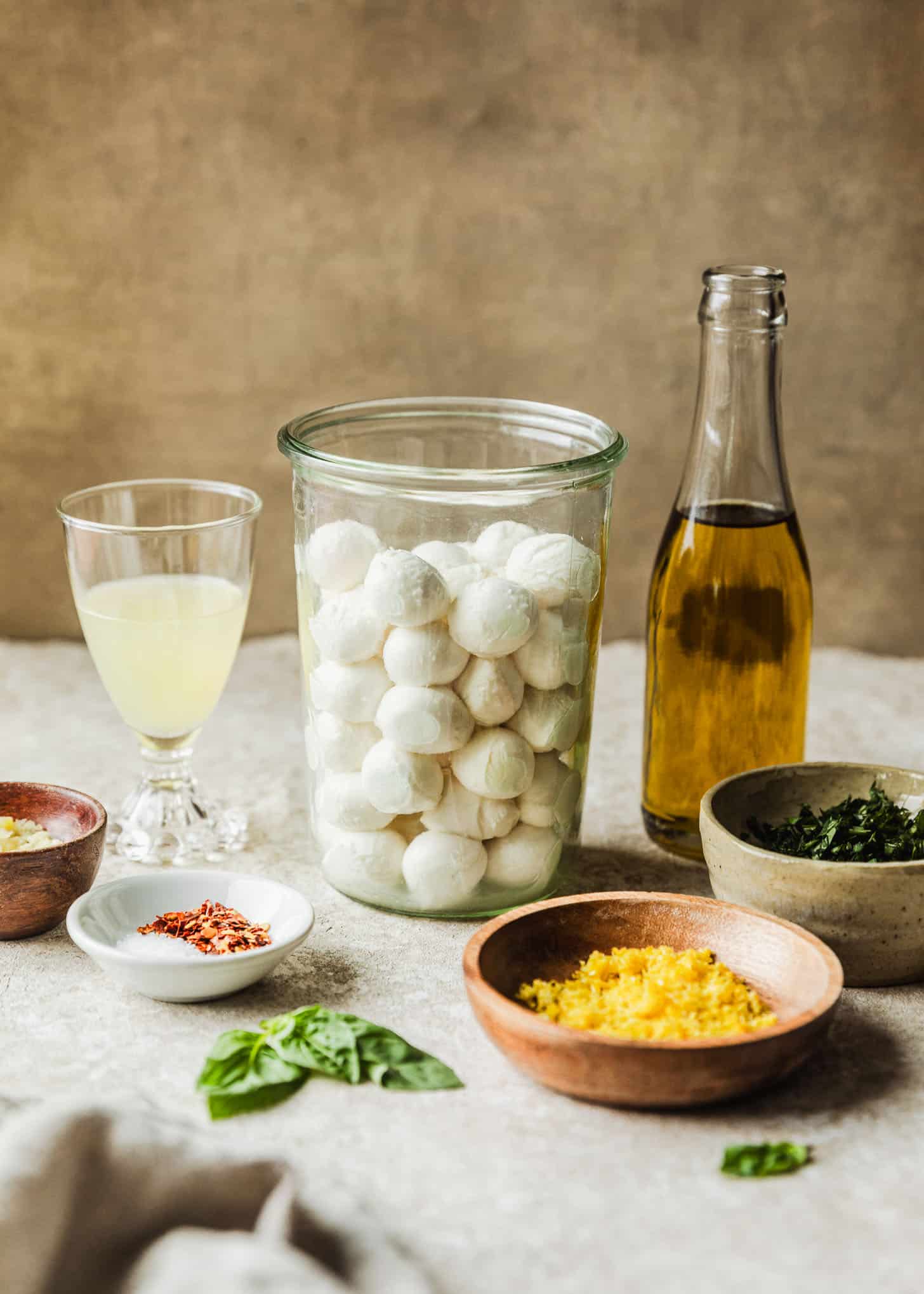 Glass jars of mozzarella pearls, olive oil, and lemon juice on a beige and brown background next to wood bowls of lemon zest and garlic, basil, and white bowls of basil and spices.