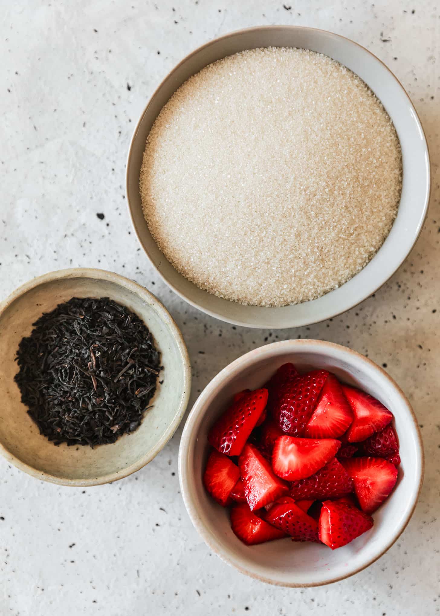 Three white bowls of sugar, black tea leaves, and strawberries on a white counter.