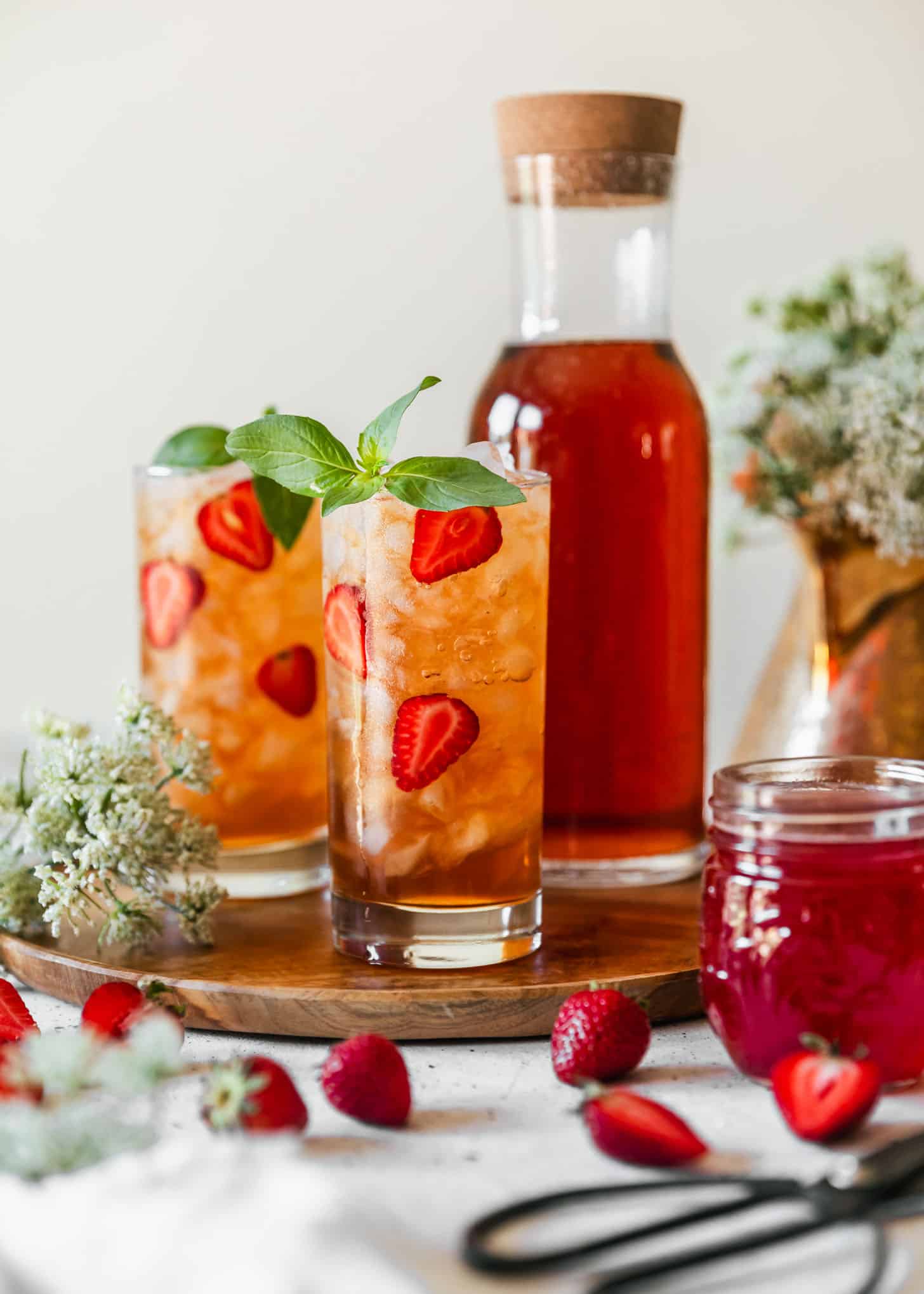 Two glasses of strawberry iced tea next to a pitcher of strawberry iced tea on a wood board next to white flowers, strawberries, a jar of strawberry syrup, and a gold teapot with a white background.