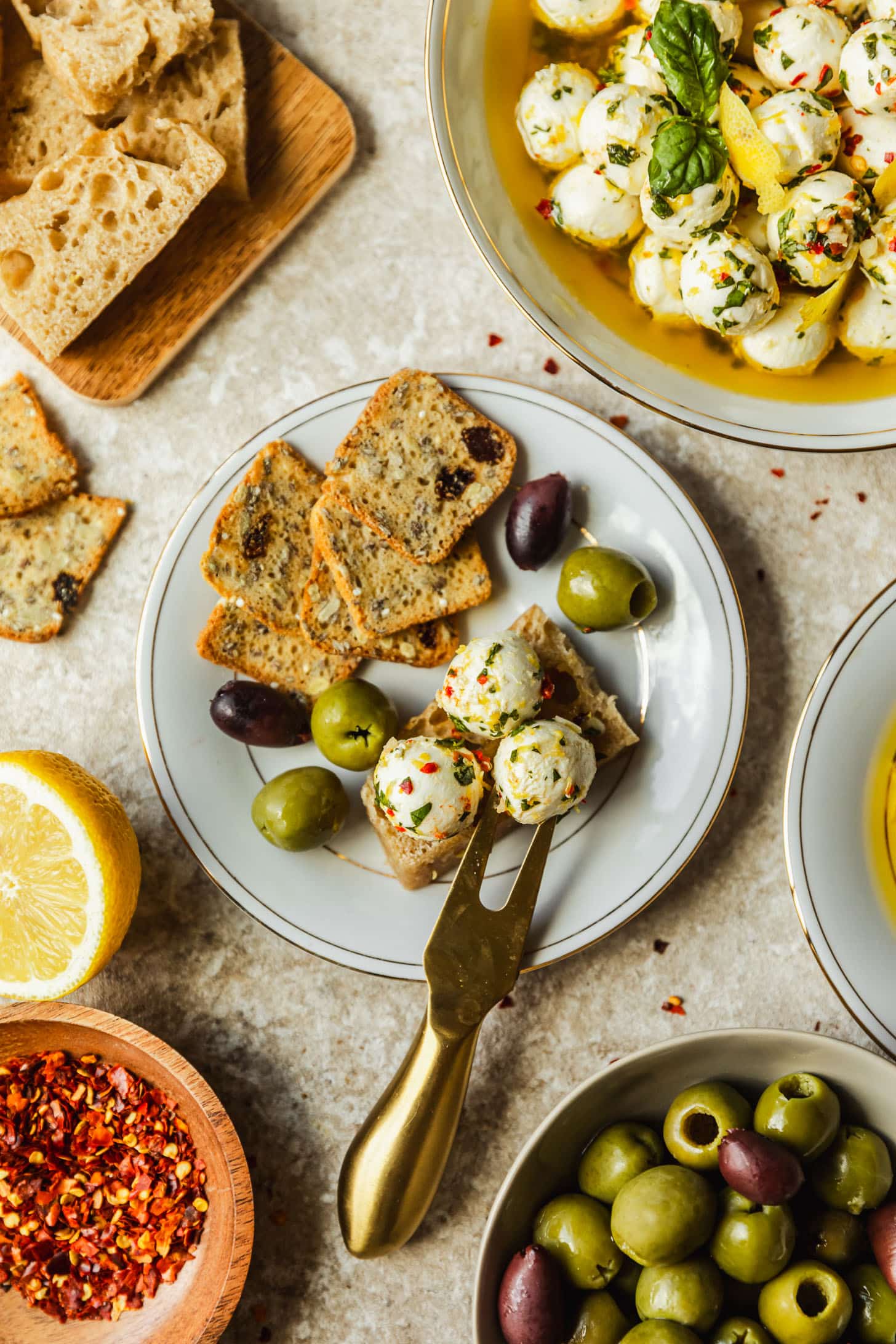 A white plate with crackers, marinated mozzarella balls, olives, and a gold cheese fork on a tan counter next to a white bowl of marinated mozzarella balls, crackers, a grey bowl of olives, a wood bowl of red pepper flakes, and a halved lemon.