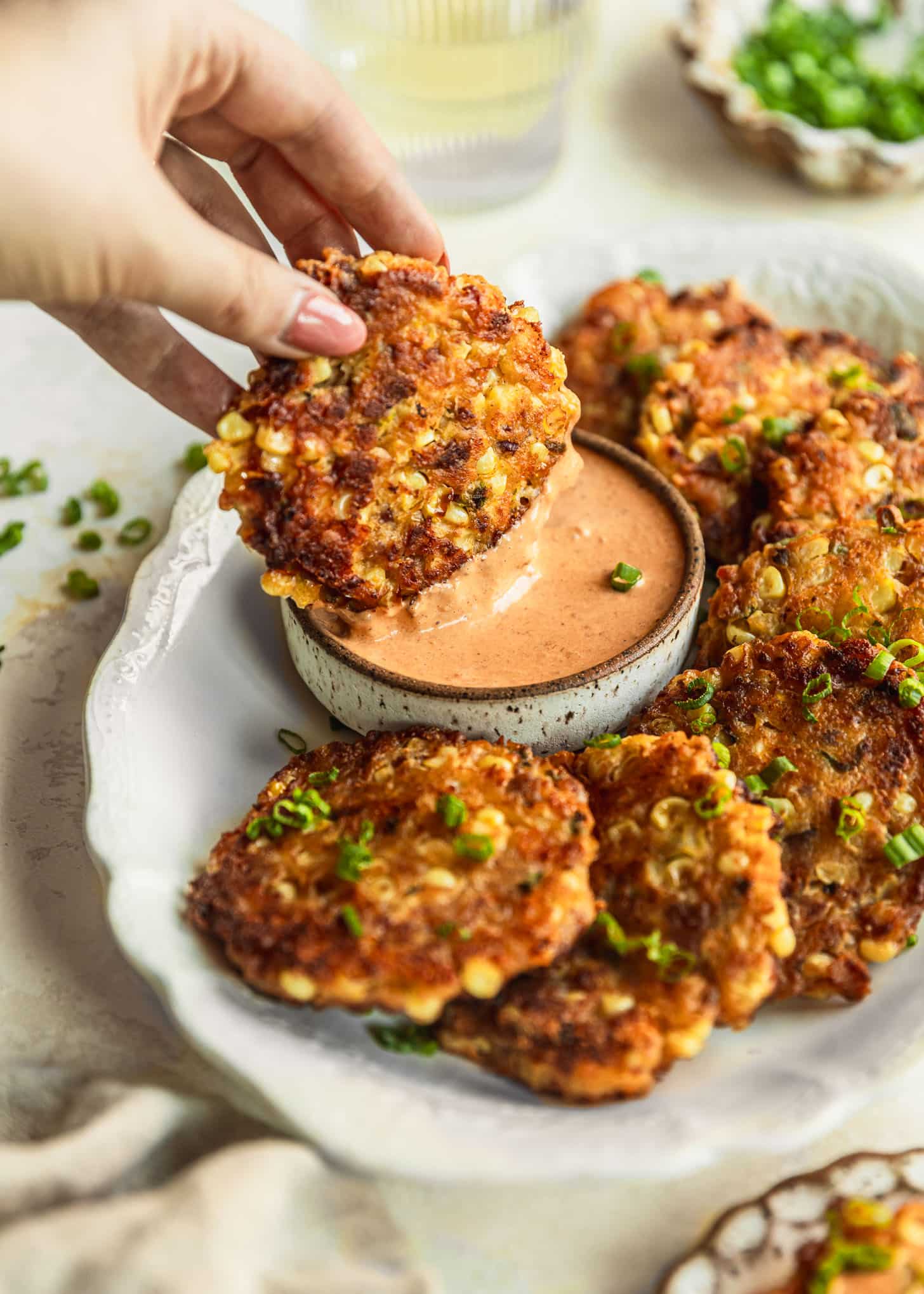 A hand dipping a fritter into a brown and white bowl of chipotle aioli on a white tray of corn and bacon fritters. The tray is on a beige counter next to a beige linen, glass of wine, and brown bowl of green onions.