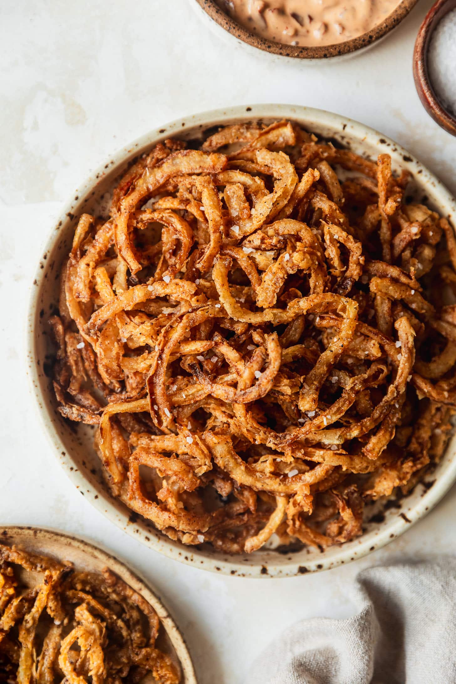 A tan speckled plate with crispy frizzled onions on a beige counter next to a tan plate of shoestring onions, wood bowl of salt, and brown bowl of chipotle aioli.