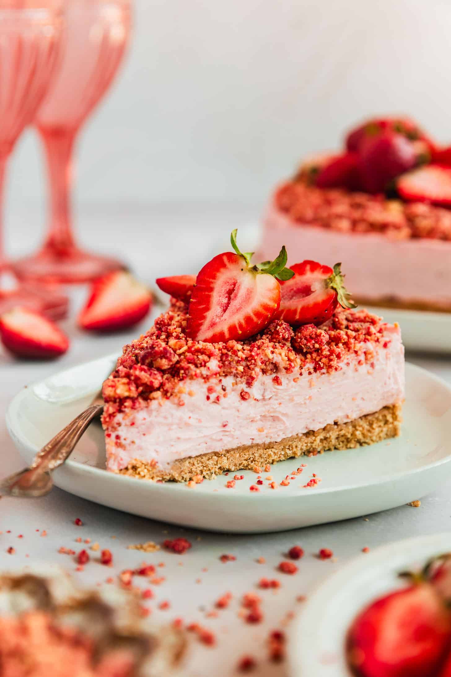 A white plate with a slice of no-bake strawberry crunch cheesecake with graham cracker crust next to a whole cheesecake, pink wine glasses, and strawberries on a pink background.