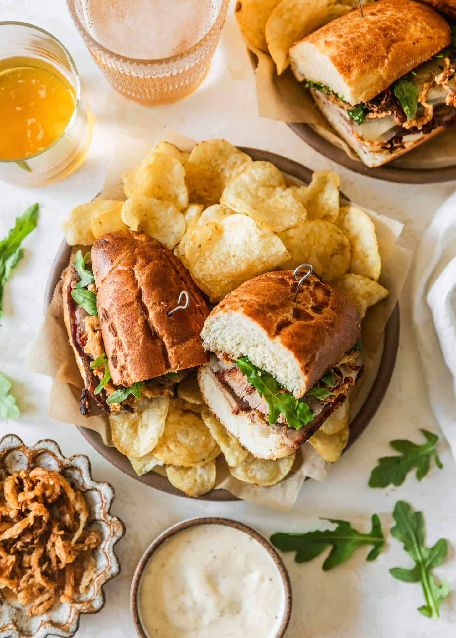 Two brown plates with tri-tip sandwiches and potato chips on a tan counter next to glasses of beer, arugula, and white and brown bowls of fried onions and garlic aioli.