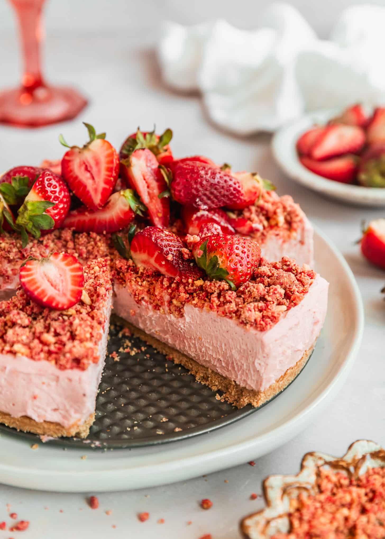 A sliced strawberry crunch cheesecake with fresh strawberries on a pink counter next to a white bowl of crunch, white bowl of strawberries, white linen, and a pink wine glass.