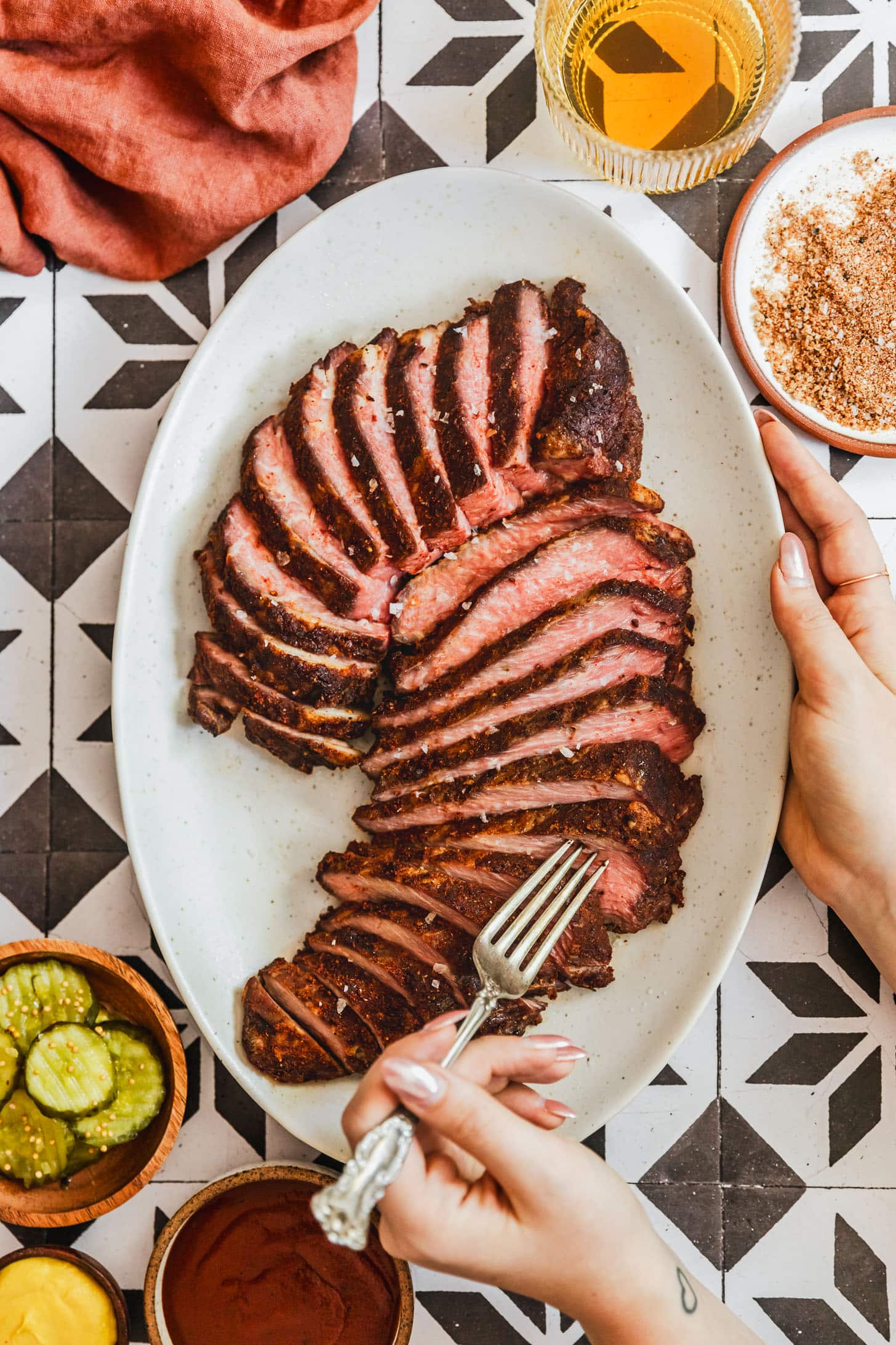 A hand grabbing a piece of smoked tri-tip with a fork off of a white platter next to a wood bowl of pickles, orange linen, glass of beer, and white bowl of BBQ rub on a black and white tile counter.