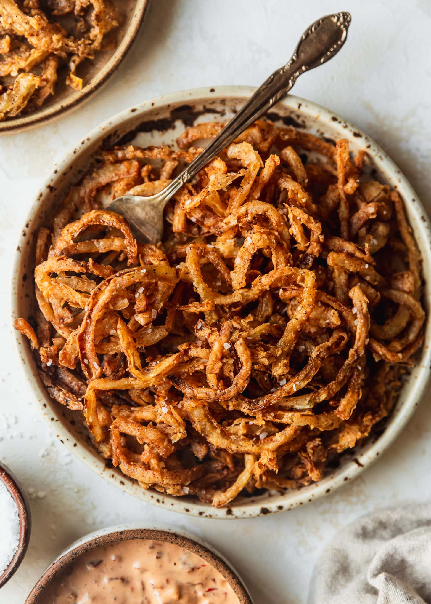 Crispy frizzled onions and a fork on white and brown speckled plate. The plate is on a beige counter next to brown bowls of salt, aioli, and onion strings.