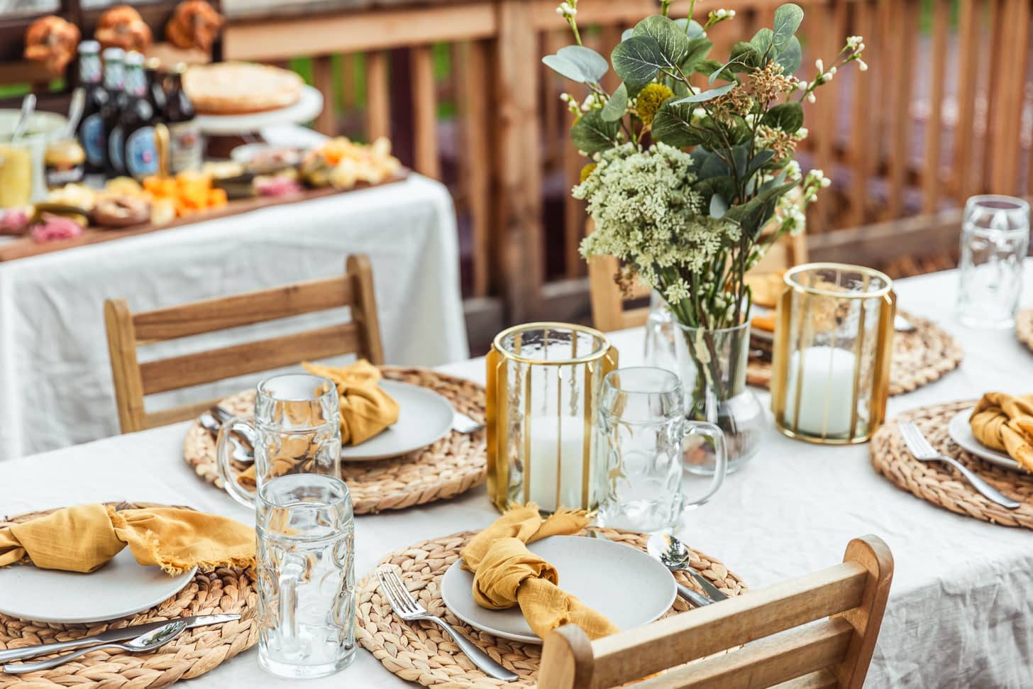A backyard Oktoberfest party with a tablescape topped with a white tablecloth, wicker placemats, yellow napkins, beer steins, and green and white flowers with a German food table in the background.