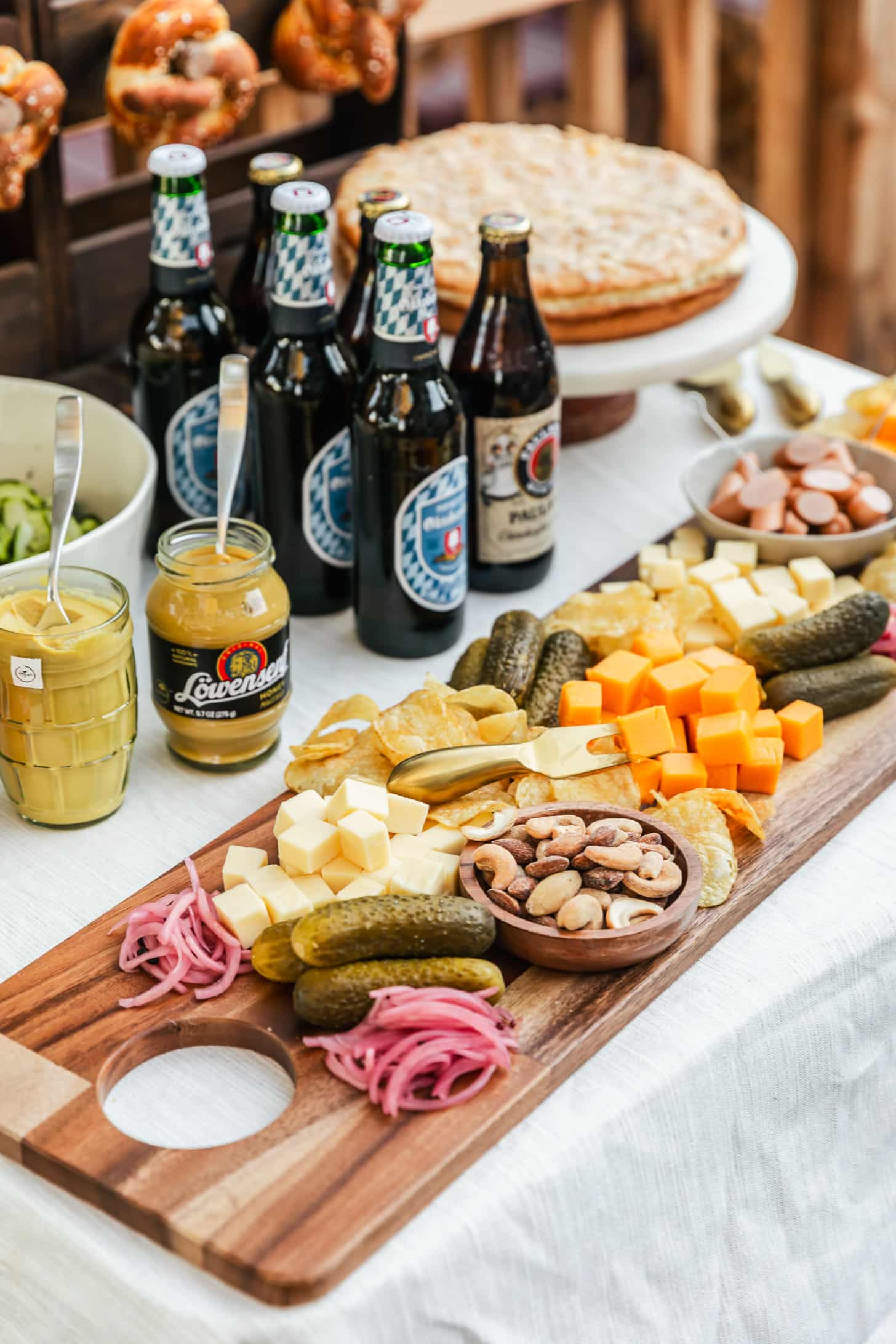 A charcuterie board, jars of mustard, German beer, and marble cake stand with an almond cake on a white tablecloth.