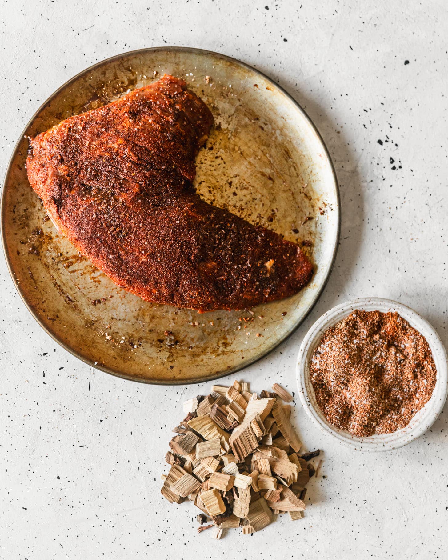 A metal tray with seasoned steak on a white counter next to wood chips and a white bowl of dry rub.