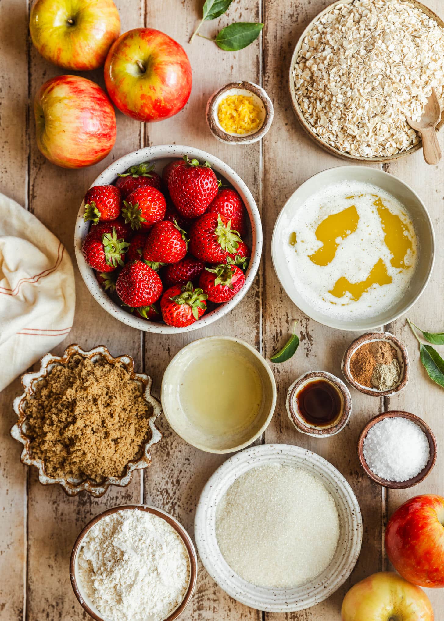 White and brown bowls of strawberries, sugar, brown sugar, lemon juice and zest, vanilla, spices, flour, and butter on a wood table.