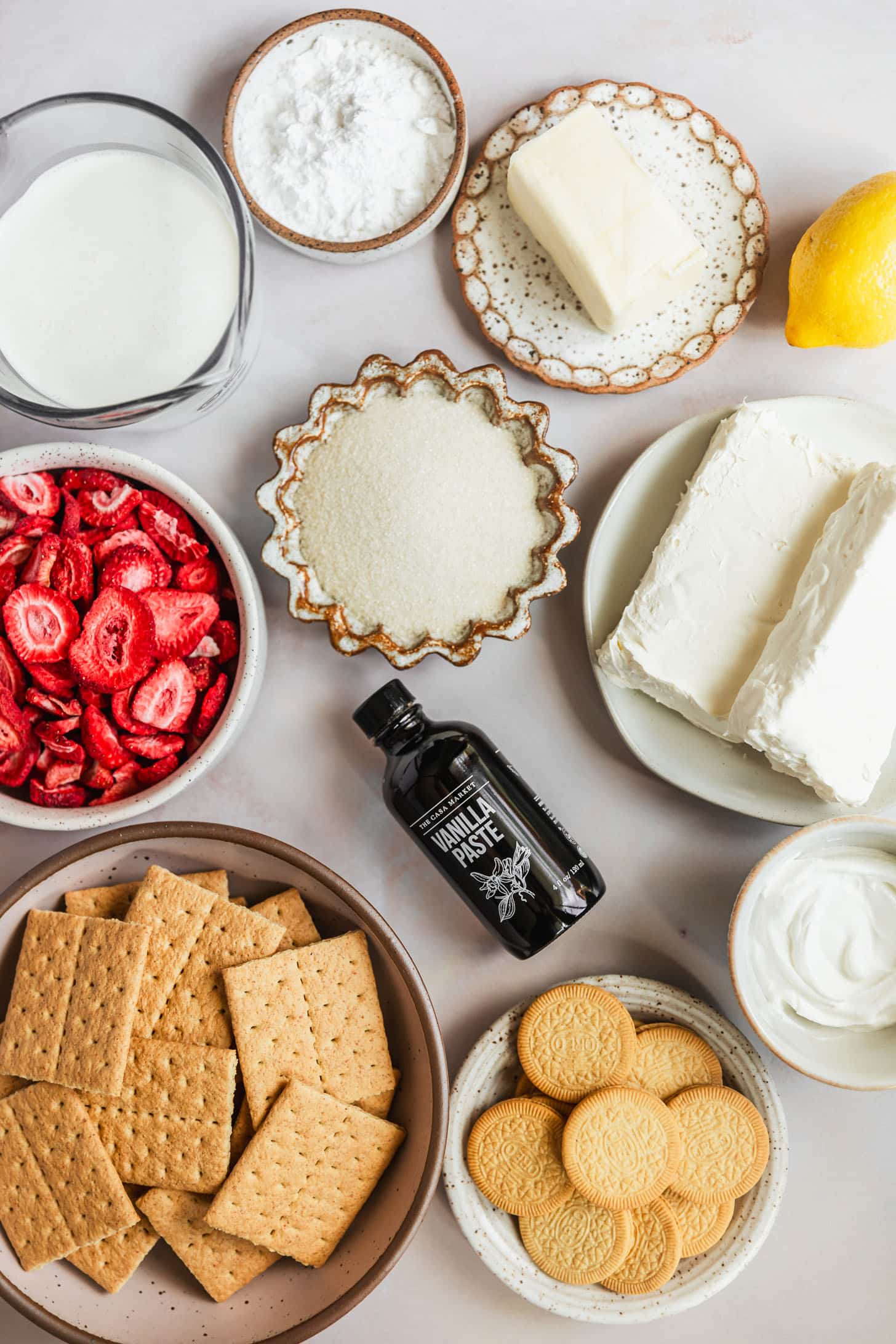 White bowls of sugar, golden Oreos, graham crackers, freeze-dried strawberries, cream cheese sour cream, butter, powdered sugar, and cream on a pink counter next to a lemon and a jar of vanilla bean paste.