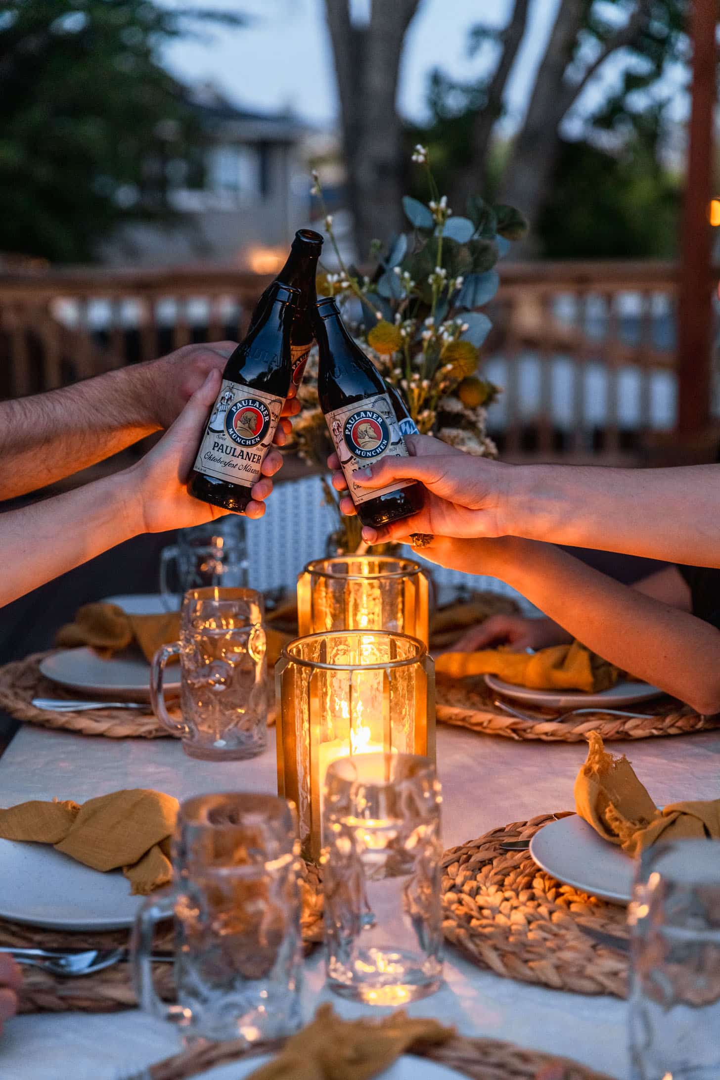 How to throw an Oktoberfest party | People doing a 'cheers' with German beer at dusk over a tablescape with a white background, candles, yellow napkins, beer steins, and wicker placemats.