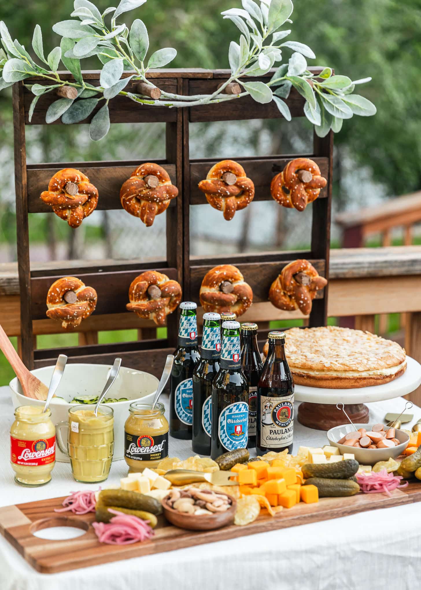 How to throw an Oktoberfest party at home | A food spread with a charcuterie board, mustard jars, beer, almonds cake, and white bowl of cucumber salad on a white tablecloth next to a board with soft pretzels.