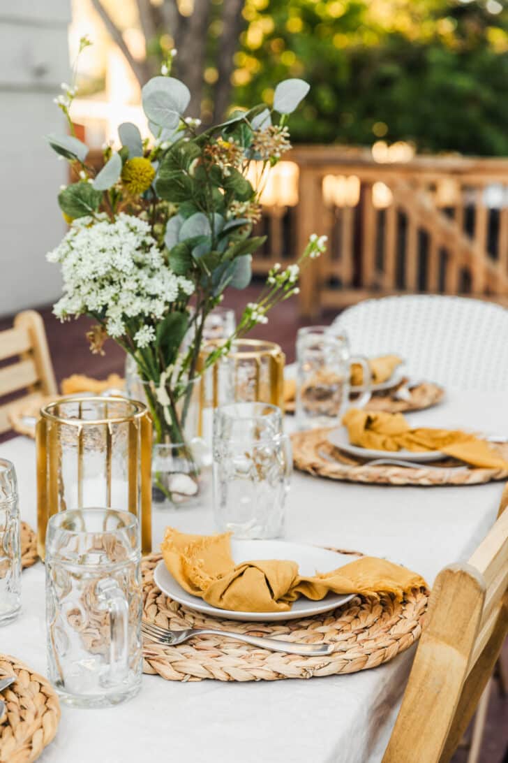 How to throw an Oktoberfest party | A tablescape with a white tablecloth, wicker placemats, yellow napkins, white plates, green and white flowers, and beer steins in a backyard.