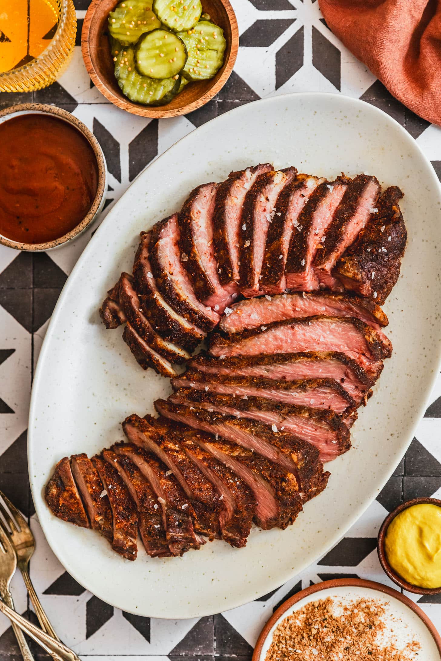 A white tray of smoked tri-tip on a black and white tile counter next to a wood bowl of pickles, brown bowl of BBQ sauce, glass of beer, orange linen, and white bowl of BBQ rub.