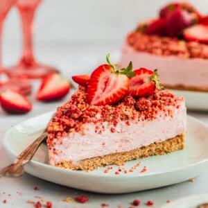 A slice of strawberry crunch cheesecake on a white plate next to a cheesecake and pink wine glasses on a pink background.