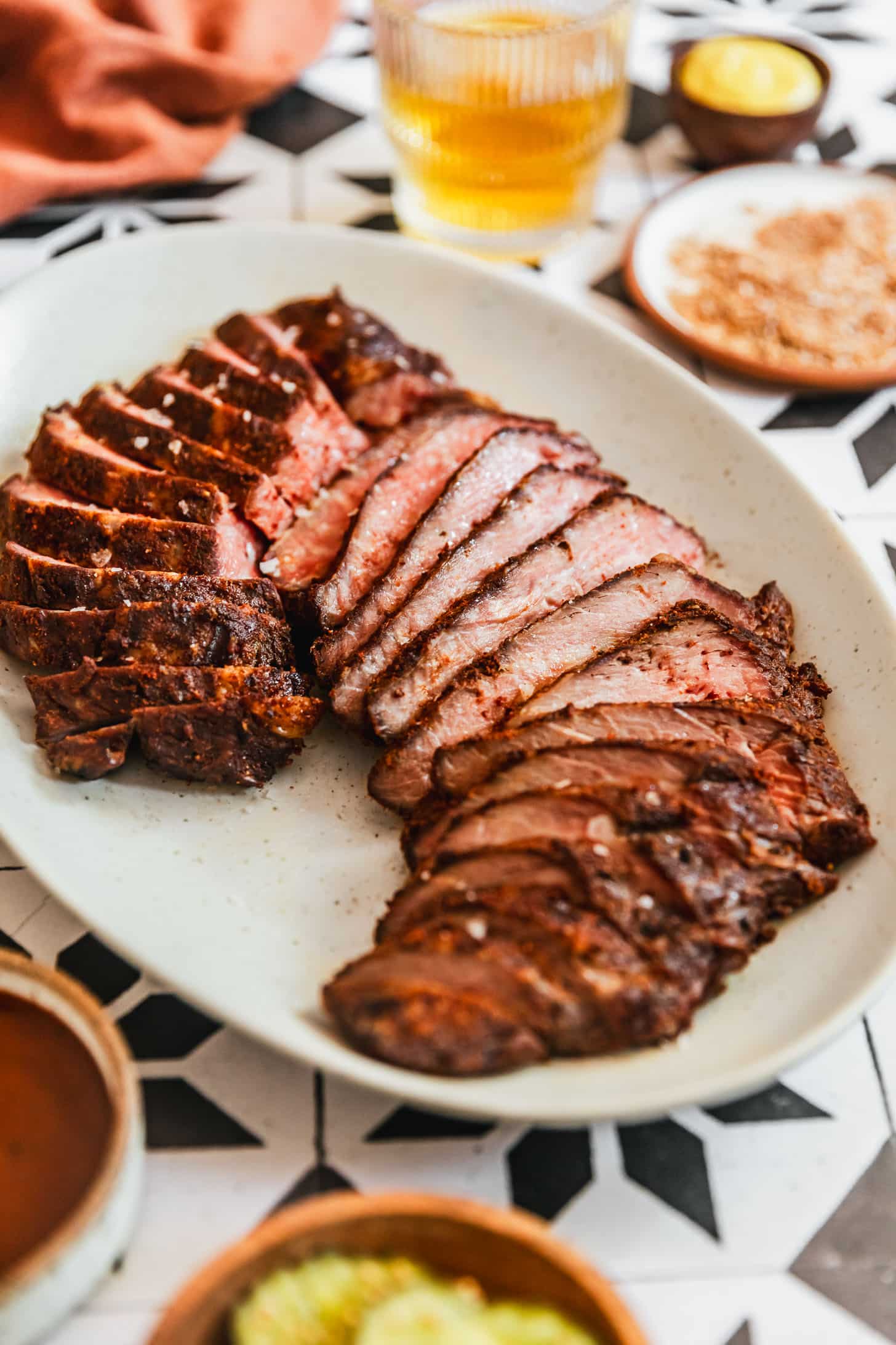 A white tray of smoked tri-tip on a black and white tile background next to a glass of beer, orange linen, white bowl of BBQ rub, and wood bowl of pickles.