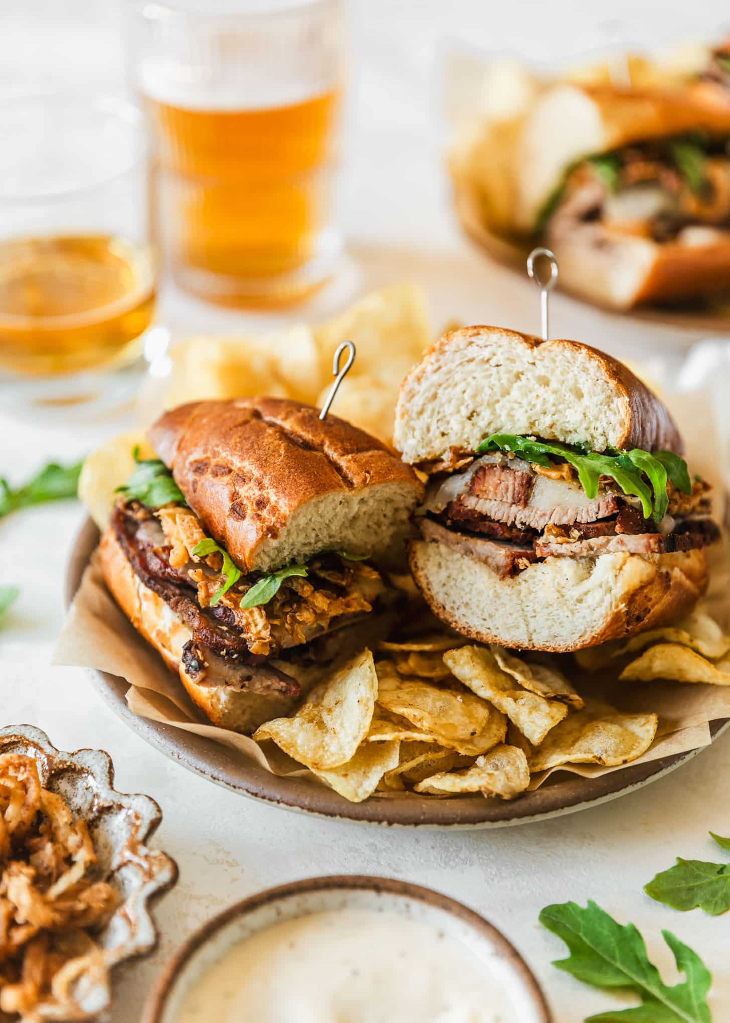 A brown plate with a tri-tip sandwich and potato chips on a beige background next to glasses of beer, a plate with a sandwich, and white and brown bowls of frizzled onions and garlic aioli.