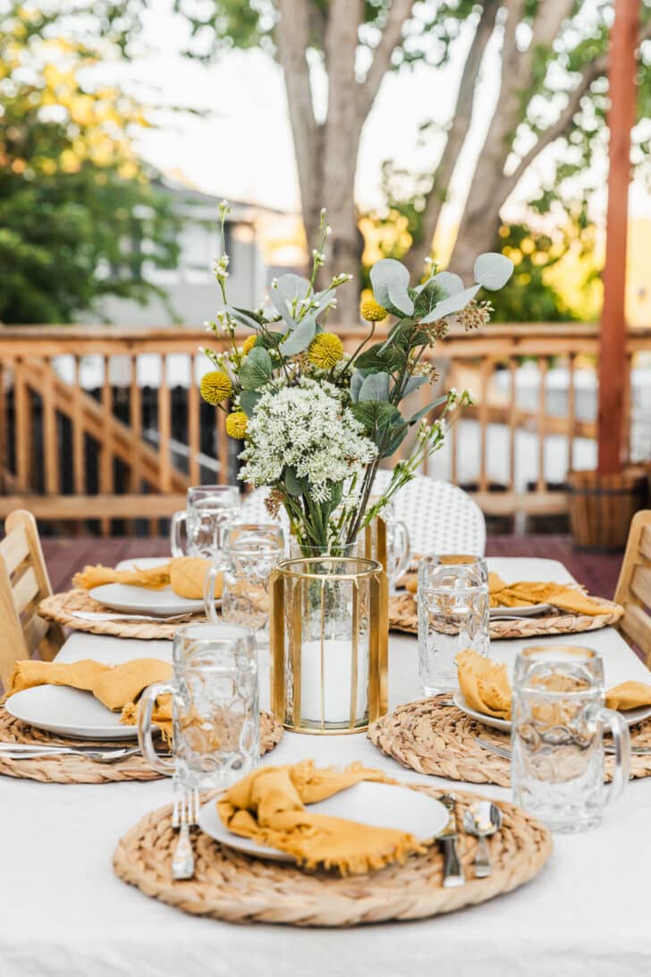 How to throw an Oktoberfest party | A DIY biergarten set up with a white tablecloth topped with place settings with wicker placemats, white plates, yellow napkins, steins, and a bouquet of green and white flowers.