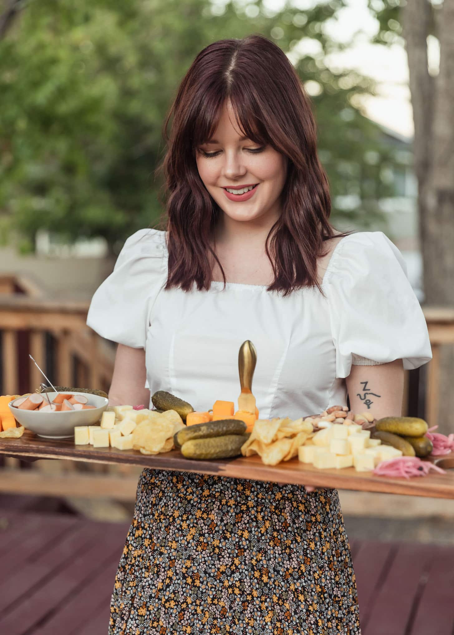 A woman with dark wavy hair and bangs wearing a white puffy shirt and black floral skirt, holding a charcuterie board, and standing in a backyard.