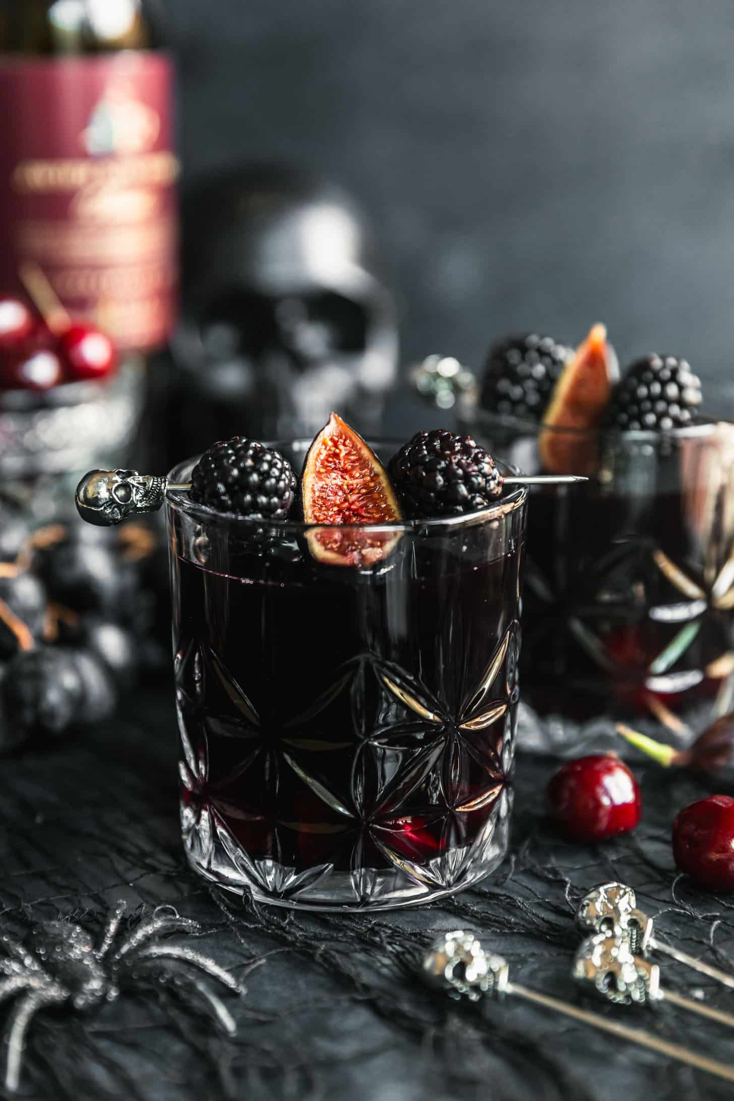 Two glasses of black Halloween sangria garnished with skull picks, blackberries, and figs on a black background next to cherries, grapes. a black skull, and a crystal glass of cherries.