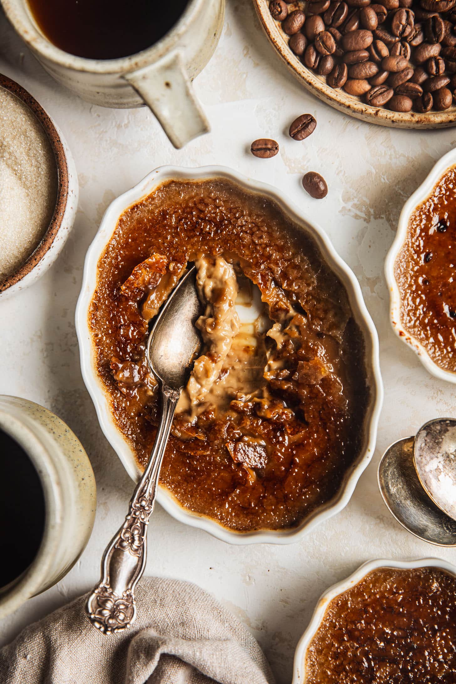 A white ramekin of coffee crème brûlée on a beige backdrop next to two more crème brûlées, two cups of coffee, a beige linen, brown bowls of coffee beans and sugar, and spoons.