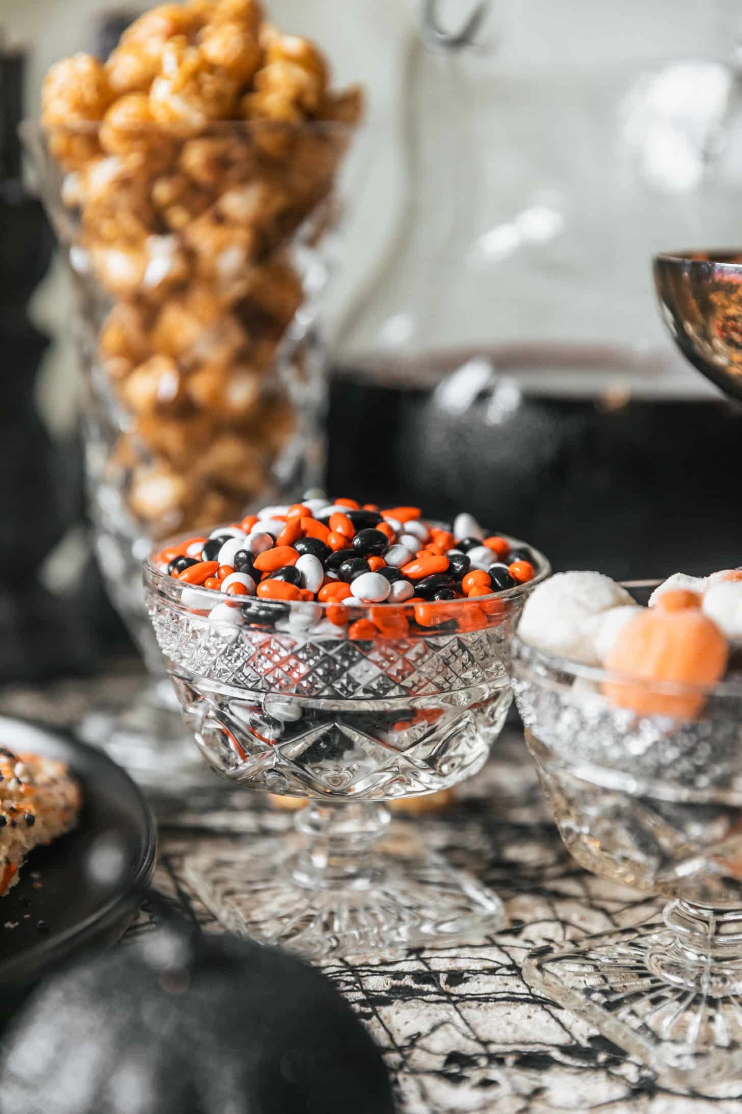 A crystal bowl of Halloween colored chocolate covered sunflower seeds on a Halloween dessert table next to a bowl of gummies, pitcher of sangria, and jar of caramel corn.