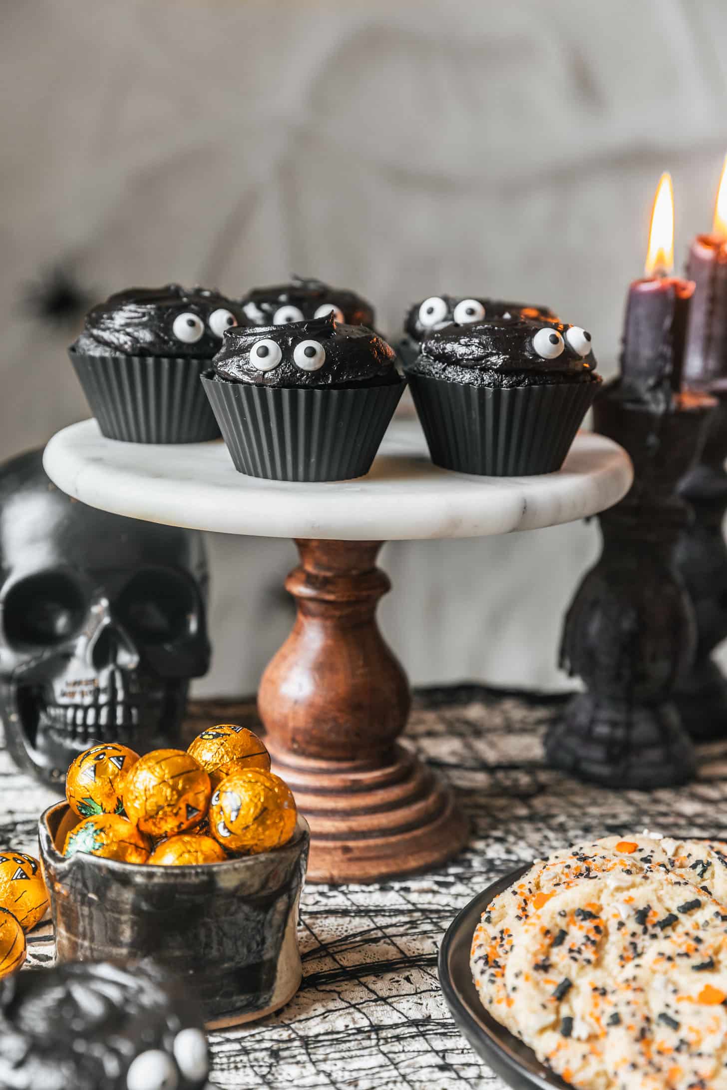 A marble cake stand with black velvet cupcakes on a black lace tablecloth next to black candles, a black skull, a black plate of cookies, and a black bowl of chocolate pumpkin candies.