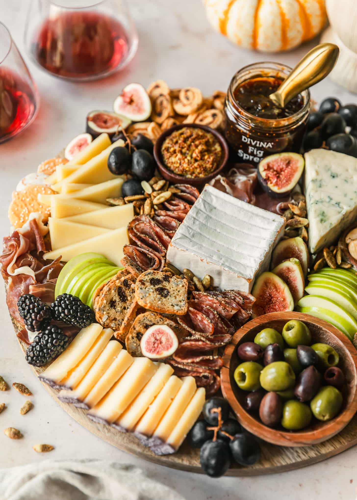 An autumn charcuterie board on a marble counter next to a beige linen, glasses of red wine, and pumpkins.