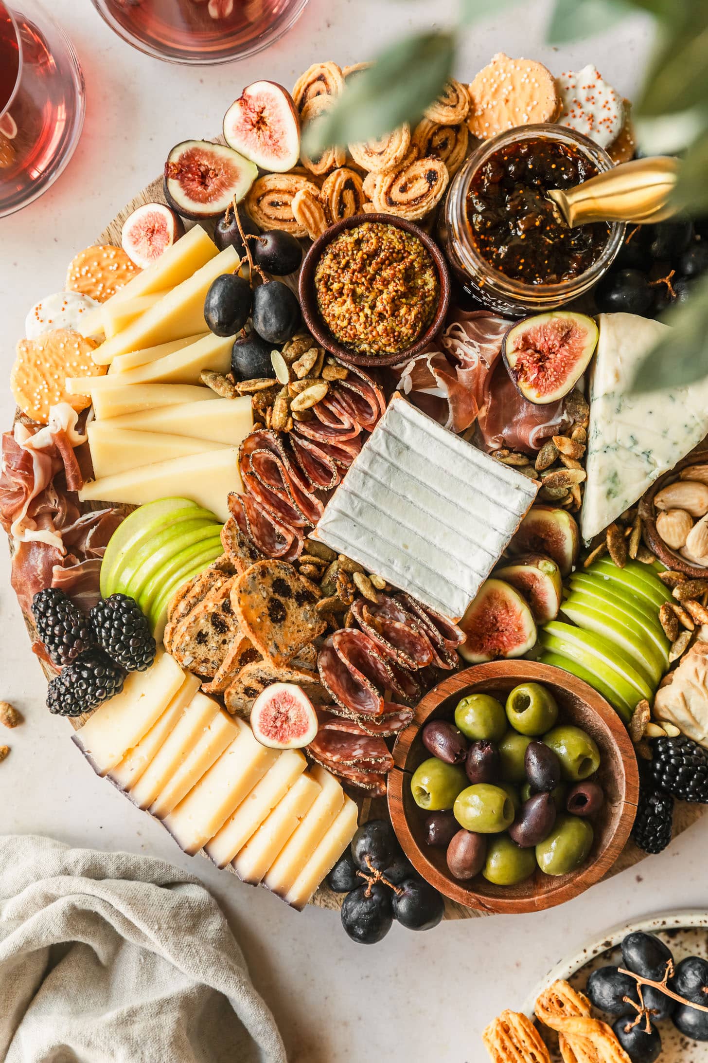 A fall charcuterie board on a beige counter next to a beige linen, glasses of red wine, greenery, and a brown plate of grapes and crackers.