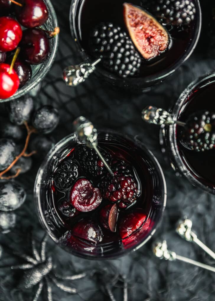 An overhead image of three glasses of black sangria with cherries, blackberries, grapes, and figs on a black counter next to grapes and plastic black spiders.