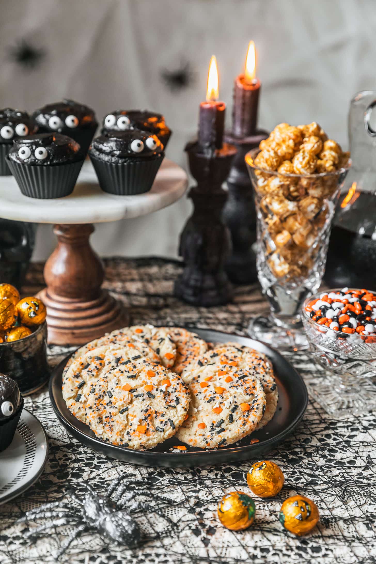 A black plate of cookies, bowls of Halloween candy, jar of caramel corn, and marble cake stand with black cupcakes on a lace tablecloth.