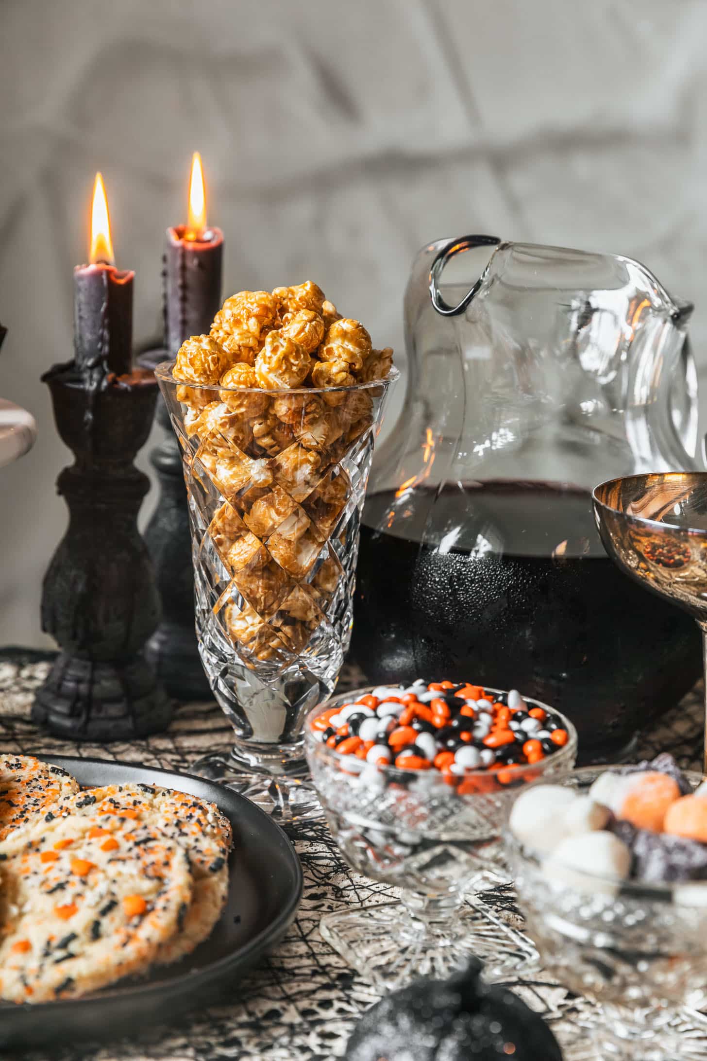 A crystal jar of caramel corn, black plate of cookies, bowls of Halloween candy, and pitcher of black sangria on a black lacy tablecloth.