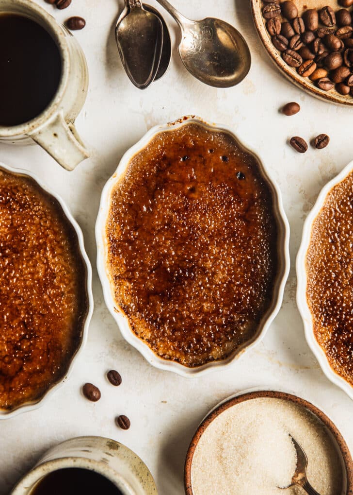 Three ramekins with coffee crème brûlée on a tan table next to spoons, two tan mugs of coffee, a brown plate of coffee beans, and a brown bowl of sugar.