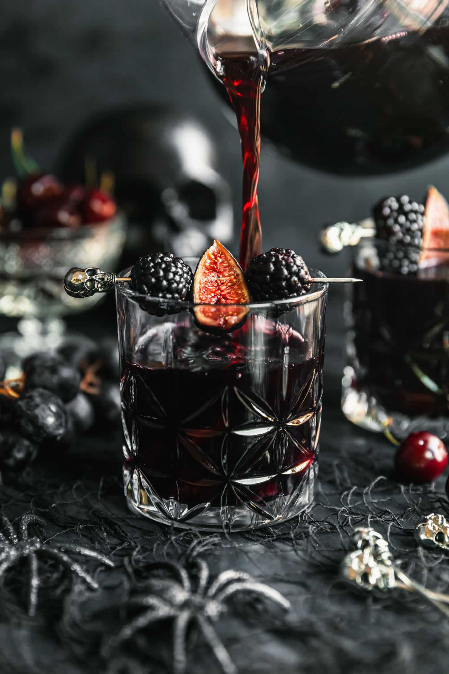 A pitcher pouring black Halloween sangria into a glass on a black counter next to grapes, cherries, plastic spiders, and a black skull.