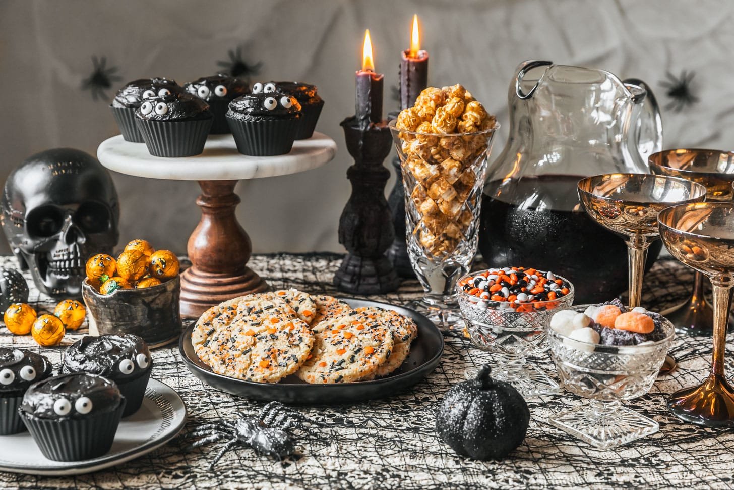 A Halloween dessert table with a lacy black table cloth topped with a black plate of cookies, marble cake stand of black velvet cupcakes, candy, a black skull, and black pumpkins.