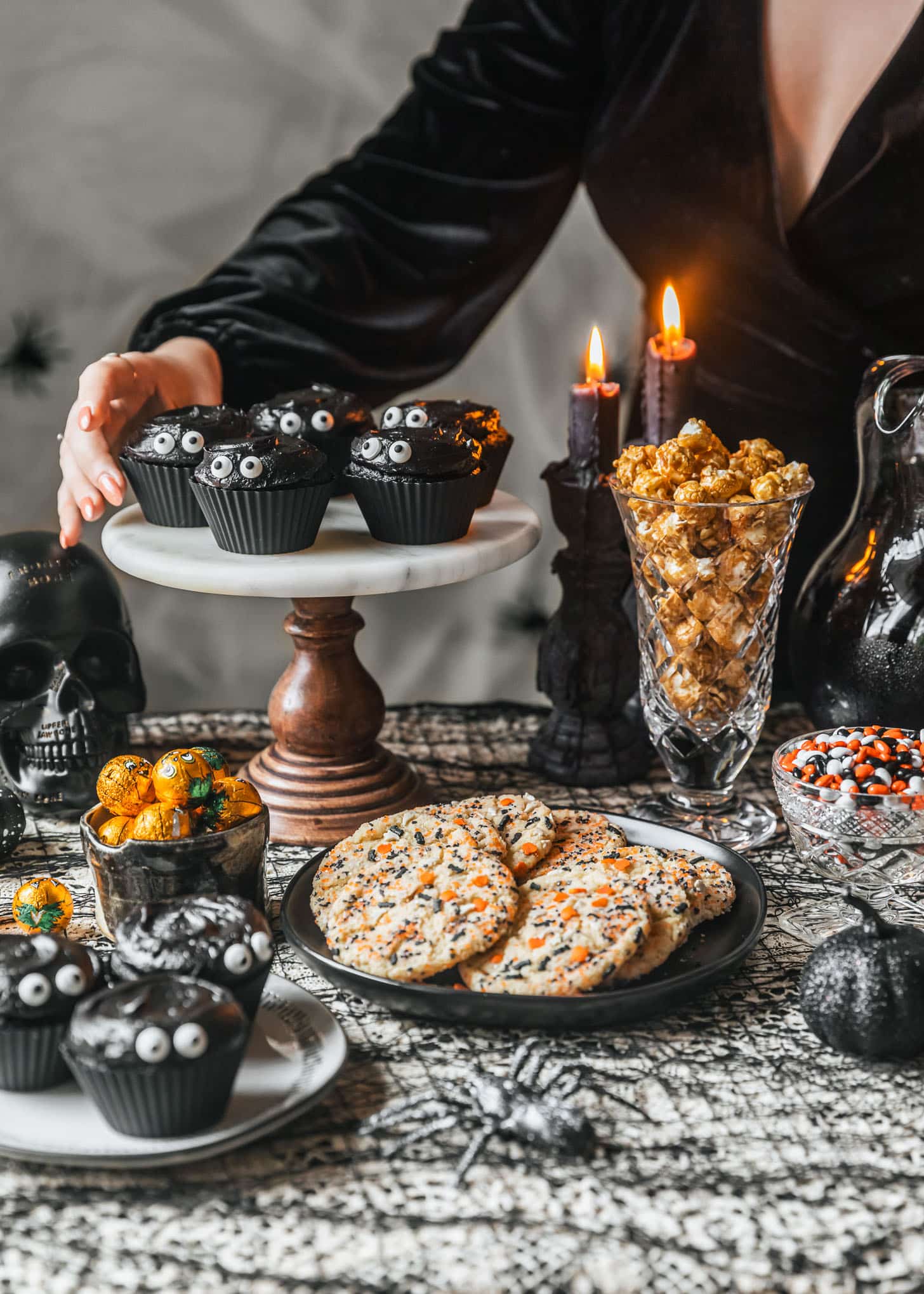 A woman in a black velvet dress grabbing a black velvet cupcake off of a marble cake stand on a Halloween dessert table next to desserts, a black skull, and black candles.