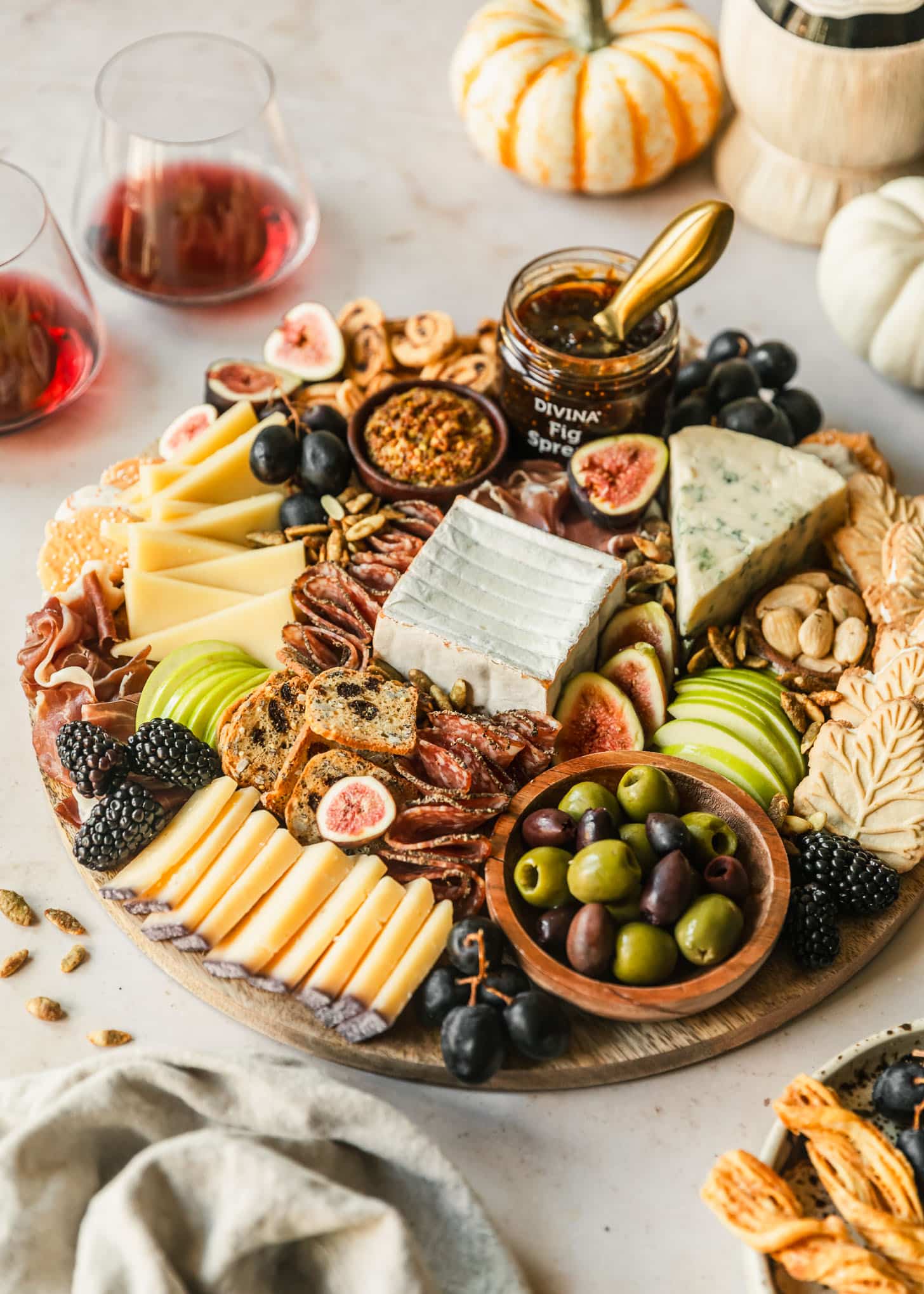 A fall charcuterie board on a marble table next to a beige linen, white plate of grapes and cheese sticks, glasses of red wine, and pumpkins.