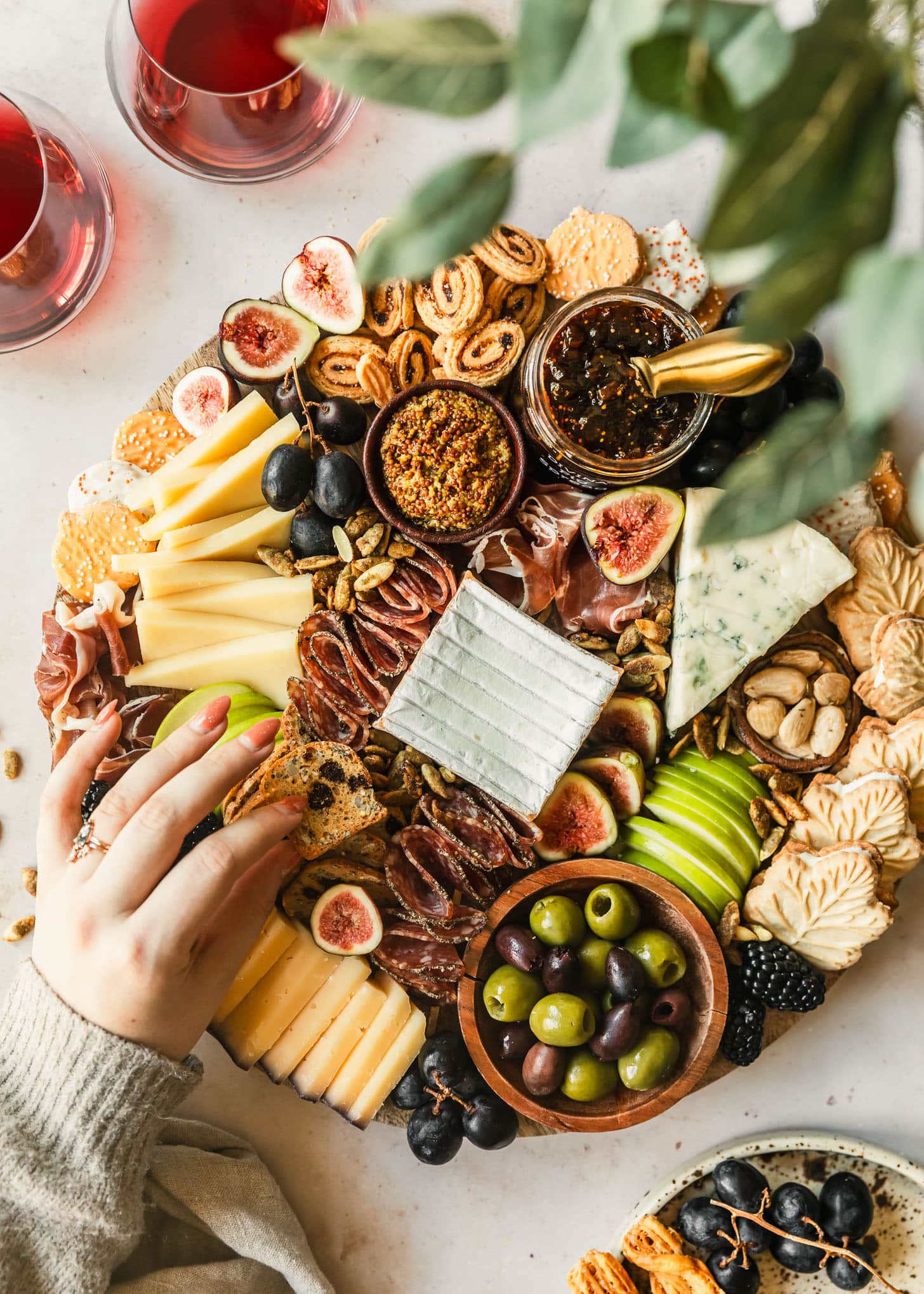 A woman's hand reaching for a cracker off of a fall charcuterie board on a tan marble counter next to a vase of greenery, glasses of red wine, and white plate of grapes.