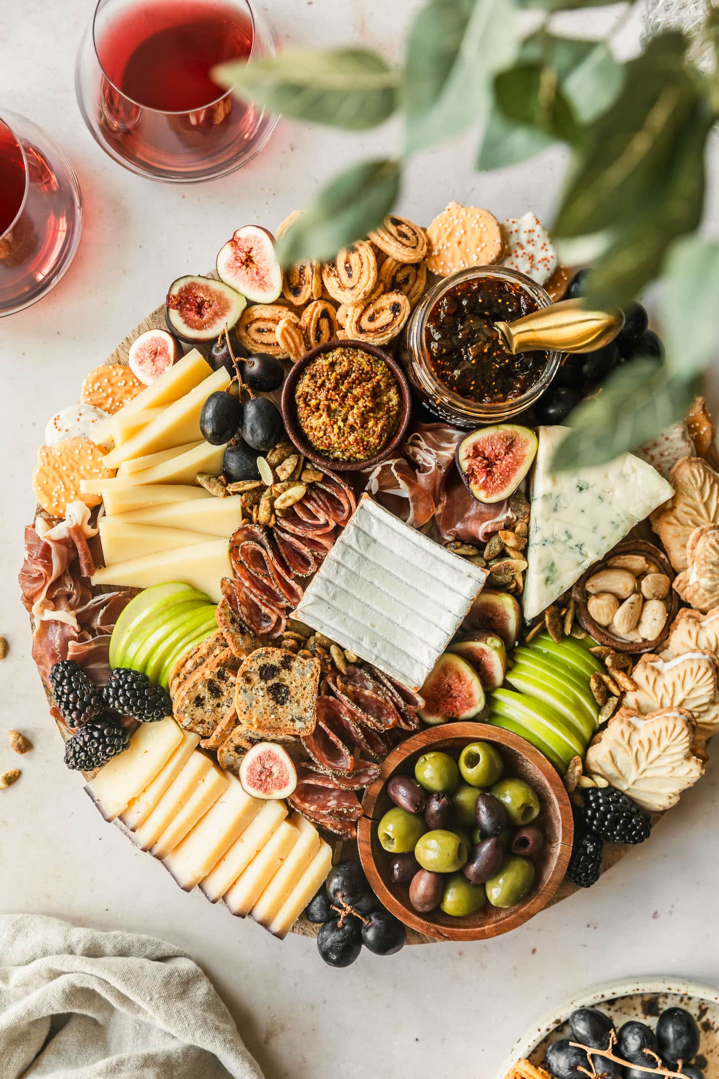 An autumn charcuterie board on a tan counter next to a beige linen, glasses of red wine, vase of greenery, and speckled plate of grapes and crackers.