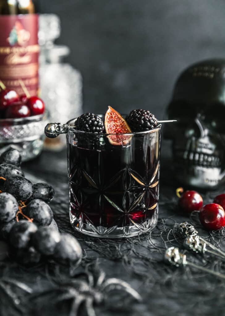 A glass of Halloween sangria garnished with blackberries and a fig on a black background next to cherries, a black skull, and a bottle of red wine.