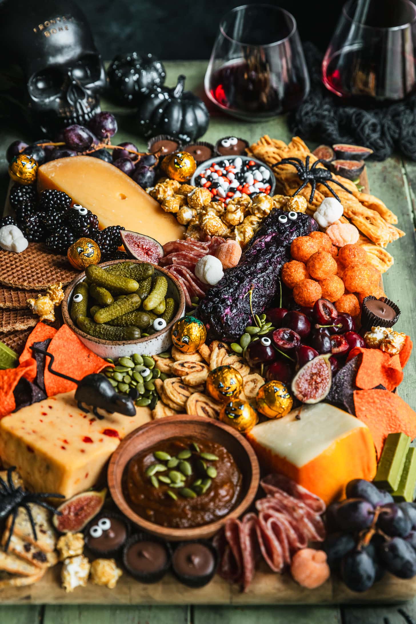 A Halloween charcuterie board on a green wood table next to a black skeleton, black pumpkins, and glasses of red wine.